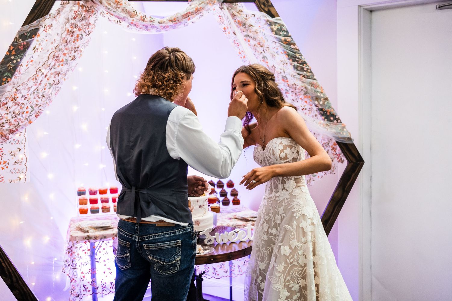 Wedding couple sharing playful moment under floral arch with string lights.