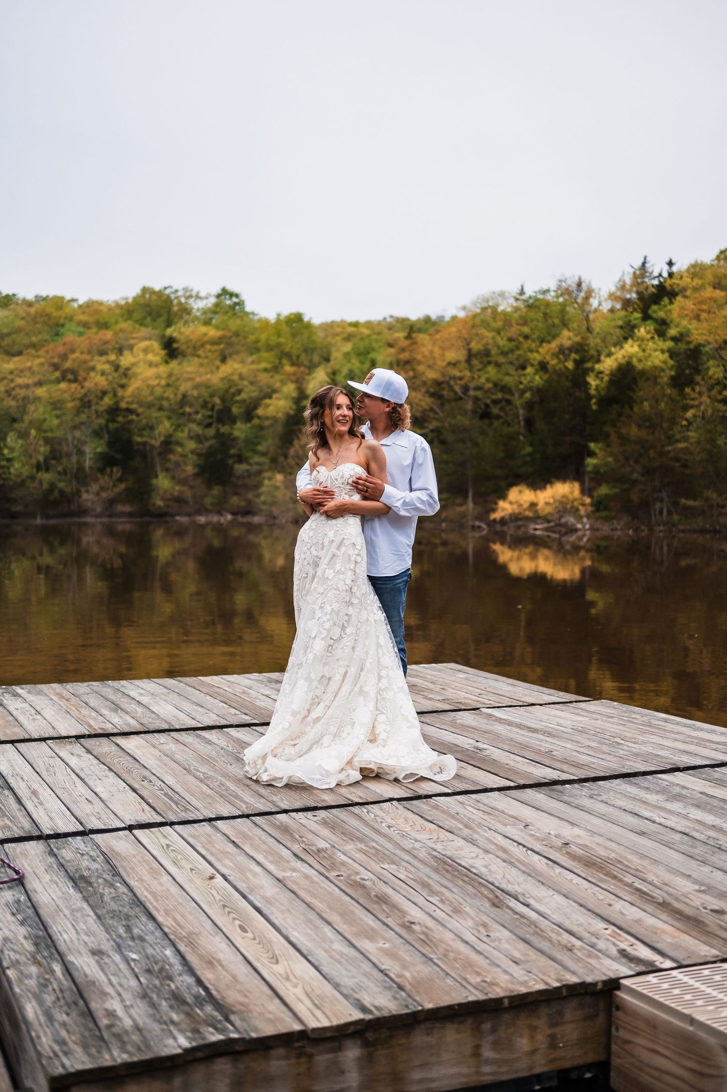 A couple embraces on a wooden dock overlooking a serene lake during an autumn wedding photoshoot.
