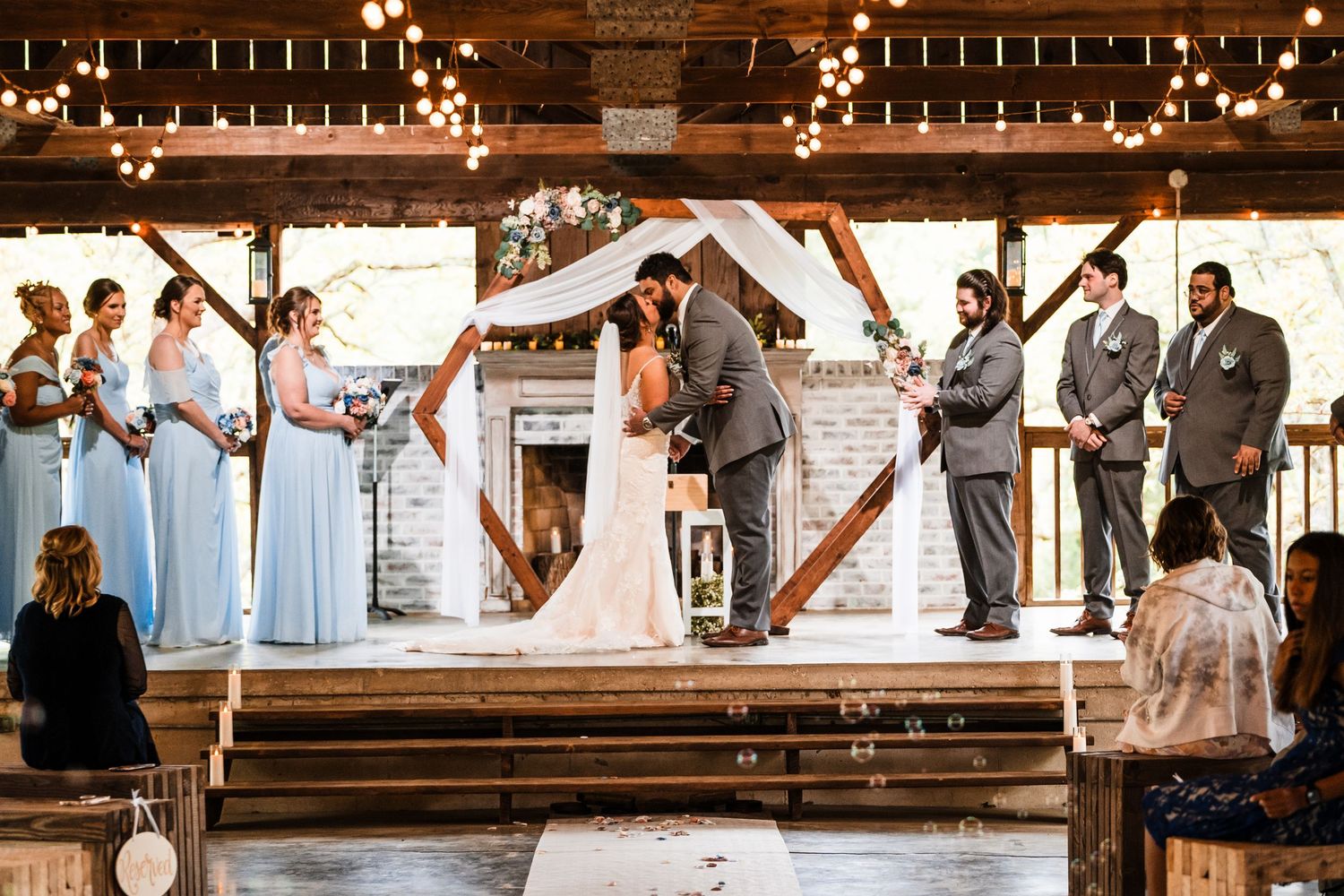 Wedding ceremony in a rustic barn venue with blue bridesmaid dresses and string lights overhead.