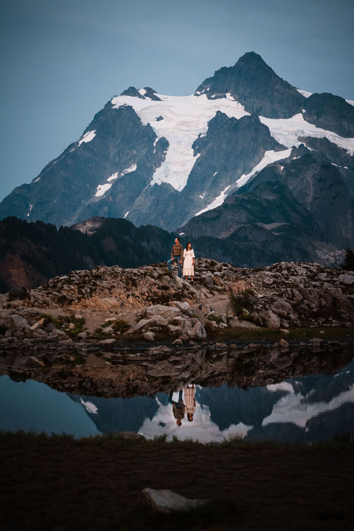 A couple stands on rocky mountain terrain with snow-capped peaks and a reflective alpine lake in the background.