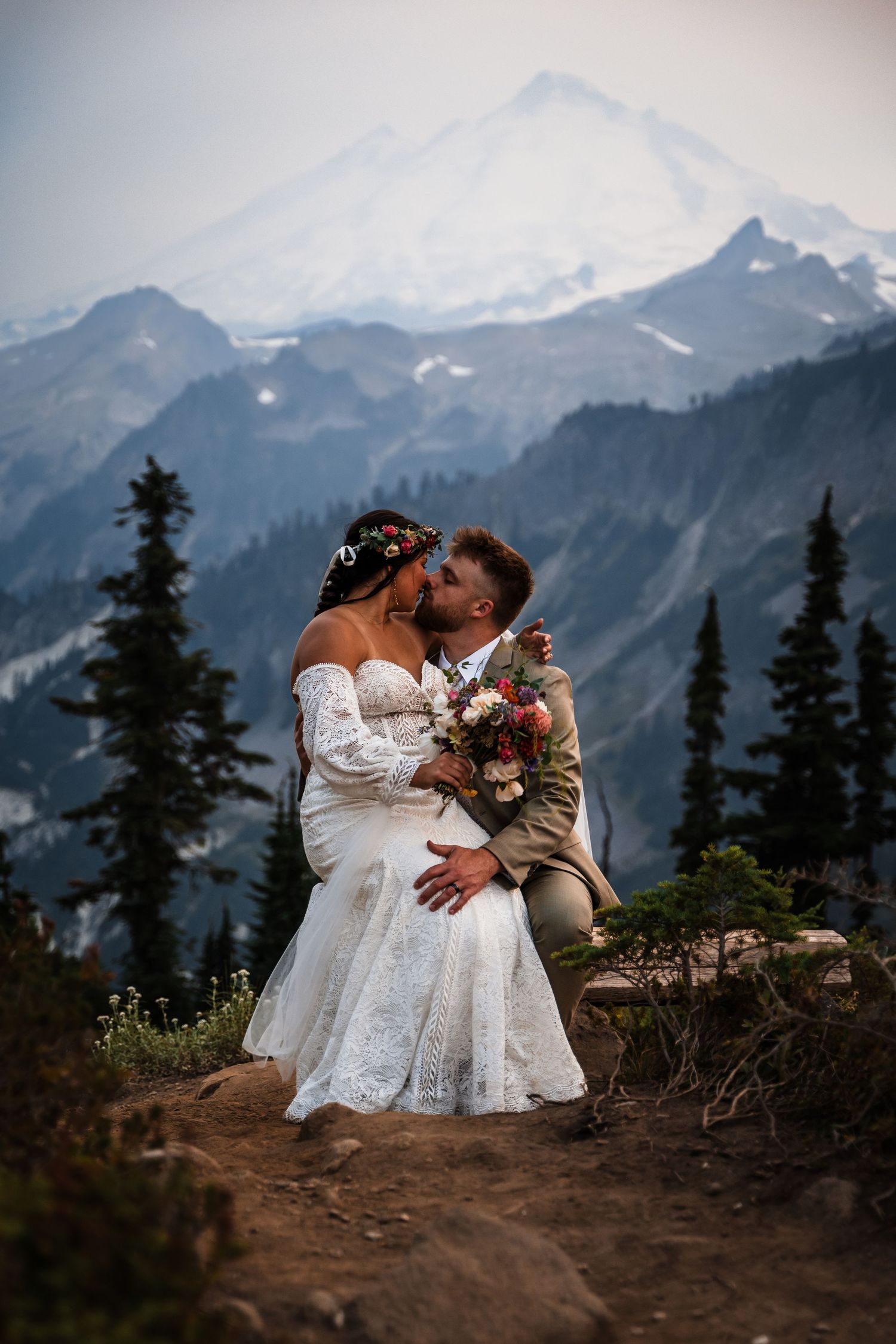 A couple in white dresses shares an intimate moment against a backdrop of majestic mountain peaks at sunset.