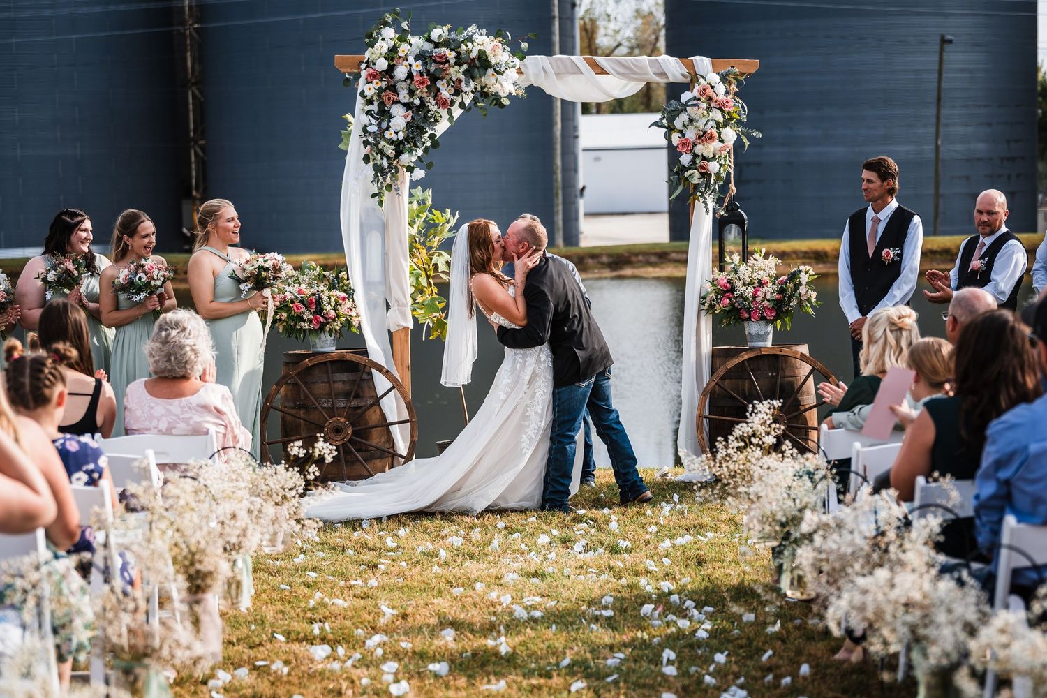 Wedding guests gather outdoors around a floral arch at a rustic blue house venue.