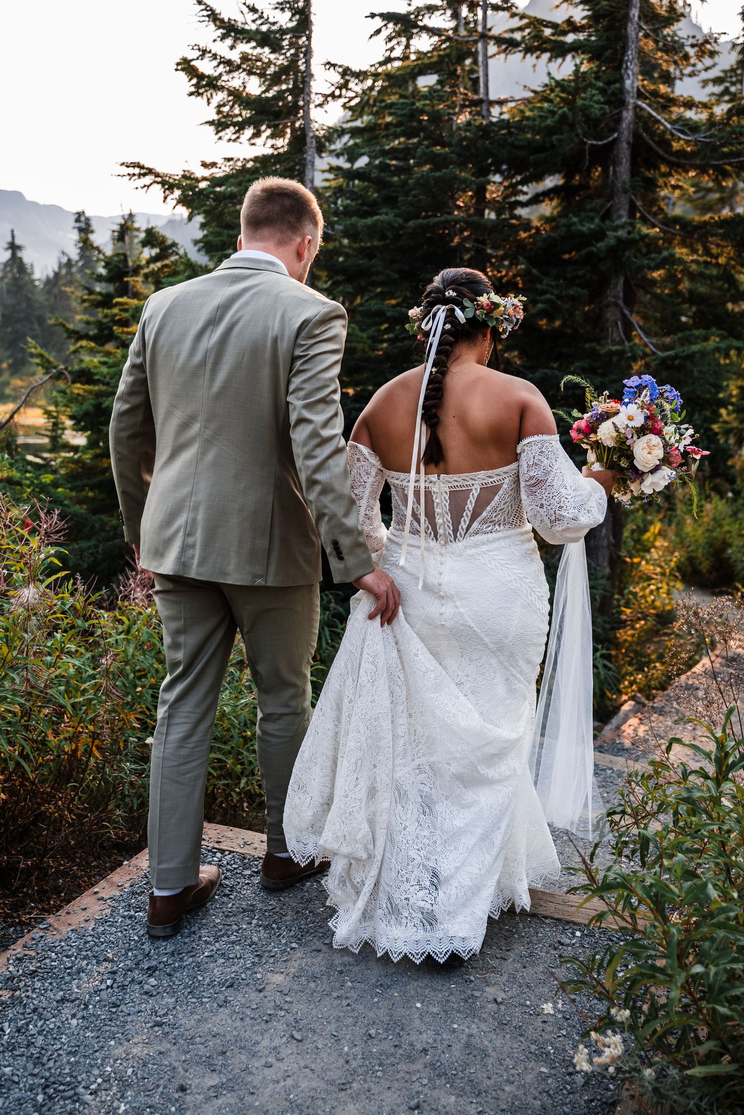 A couple in wedding attire walk along a gravel path surrounded by evergreen trees in a mountain setting.