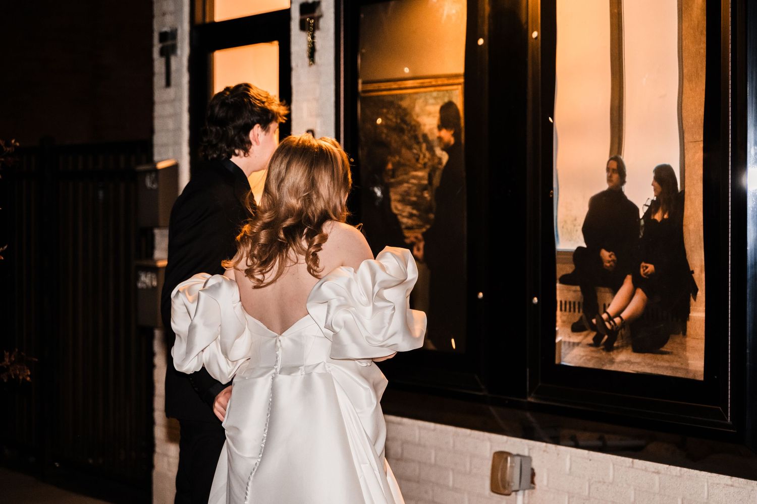 Couple in formal wear sharing an intimate moment outside a warmly lit building entrance.