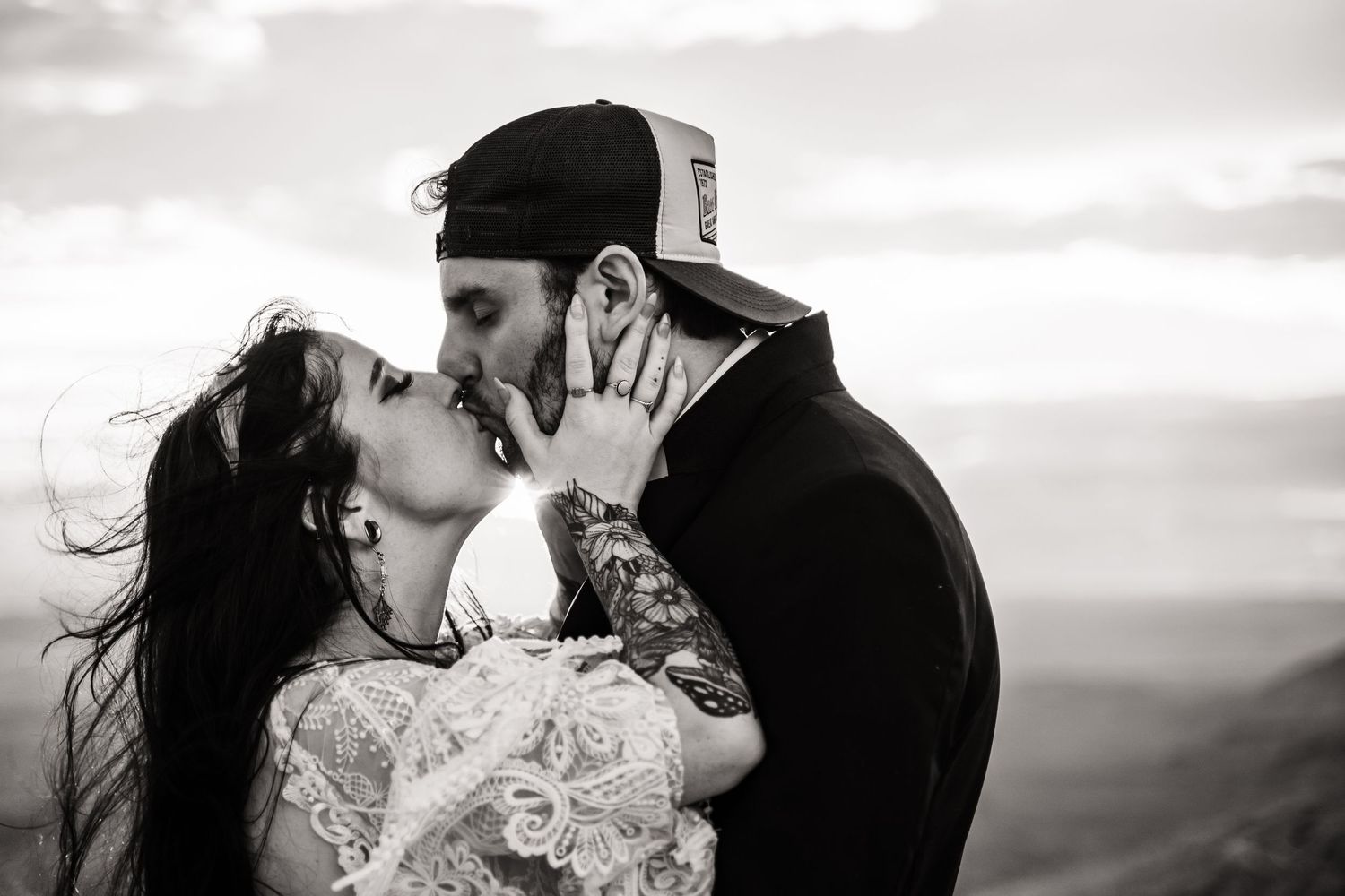 A dramatic black and white portrait captures an intimate kiss against a cloudy sky.