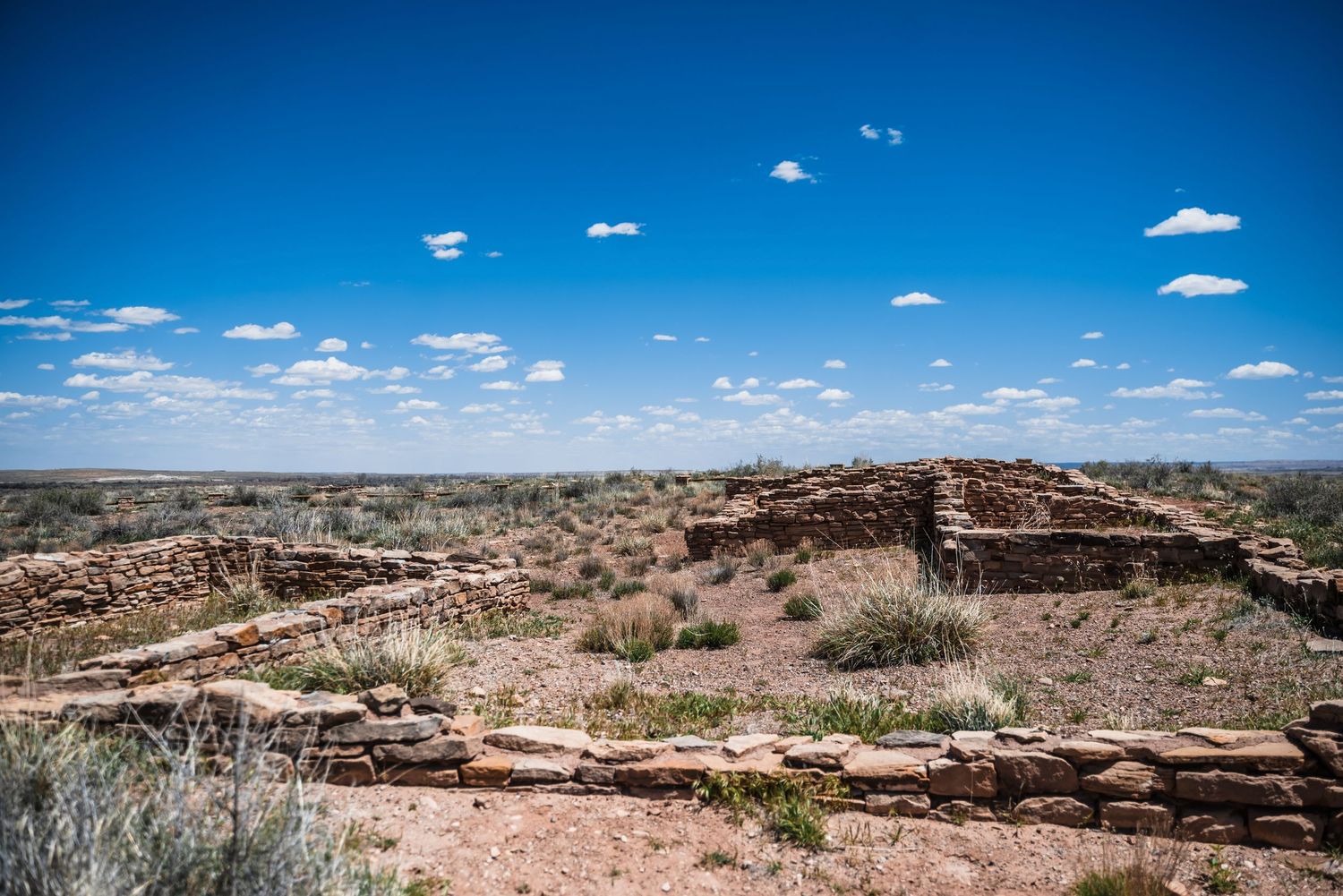 Ancient stone ruins perch atop a desert mesa under scattered white clouds and brilliant blue skies in the American Southwest.