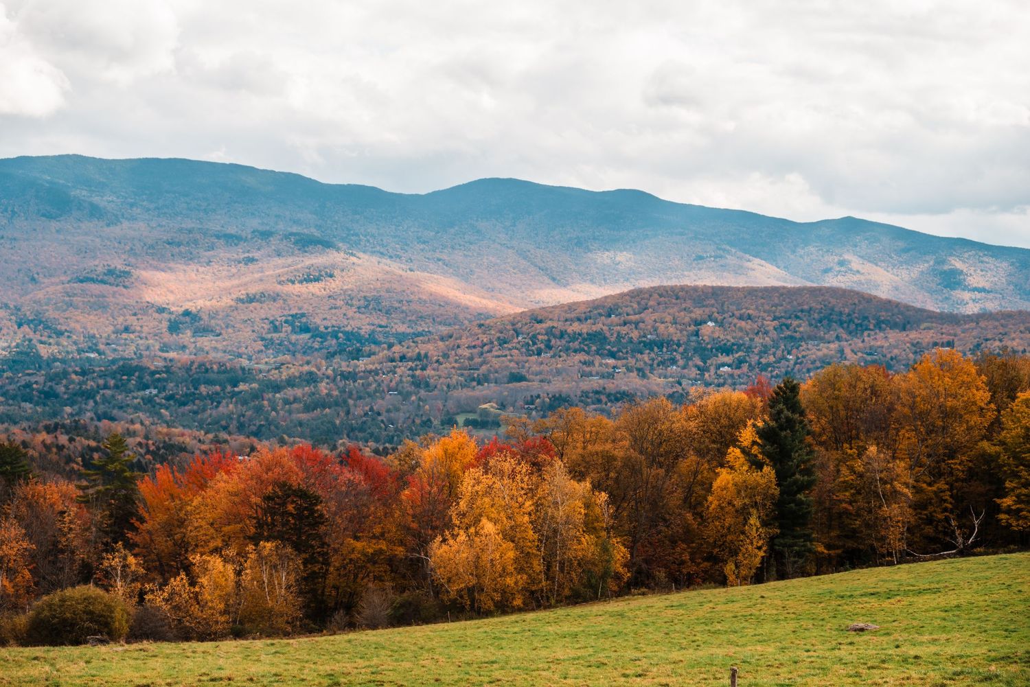 Fall foliage displays vibrant orange and yellow colors across rolling hills in the Vermont mountains under cloudy skies.