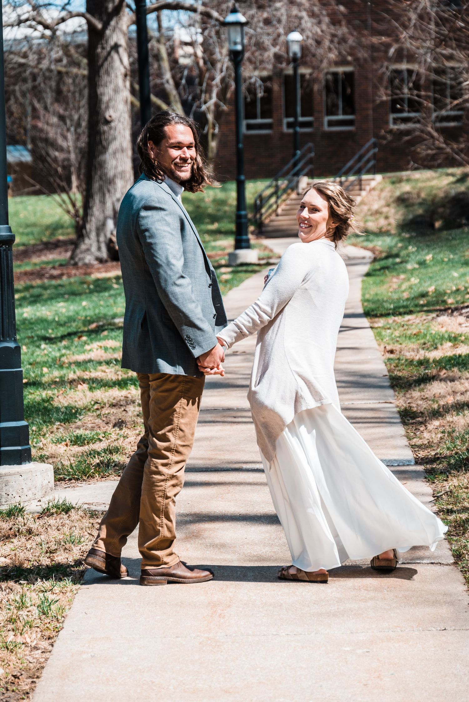 A couple in wedding attire holding hands on a campus walkway surrounded by brick buildings.
