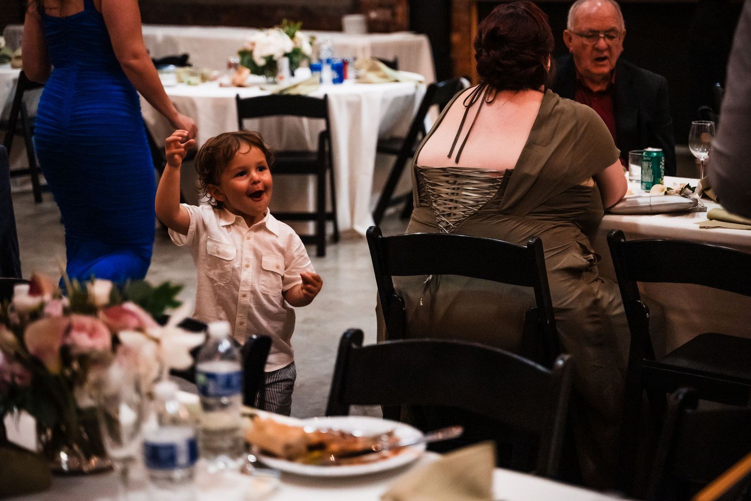 A young child in formal attire enjoying the wedding reception among dining tables.