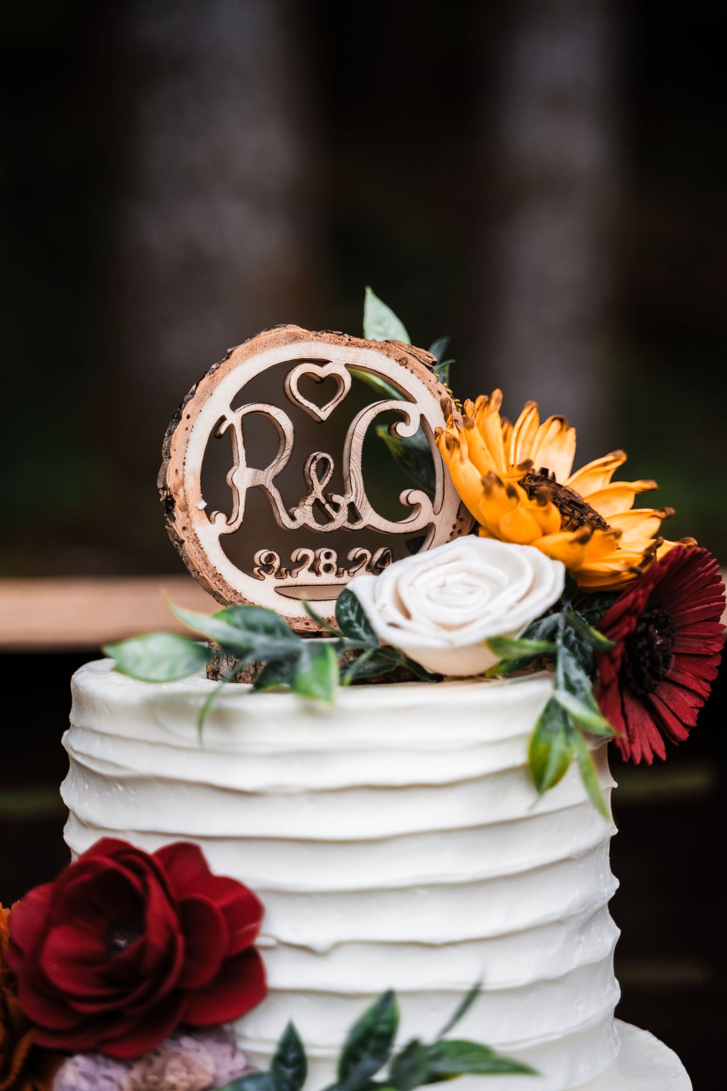 White wedding cake decorated with sunflowers, roses and a rustic wooden monogram topper.