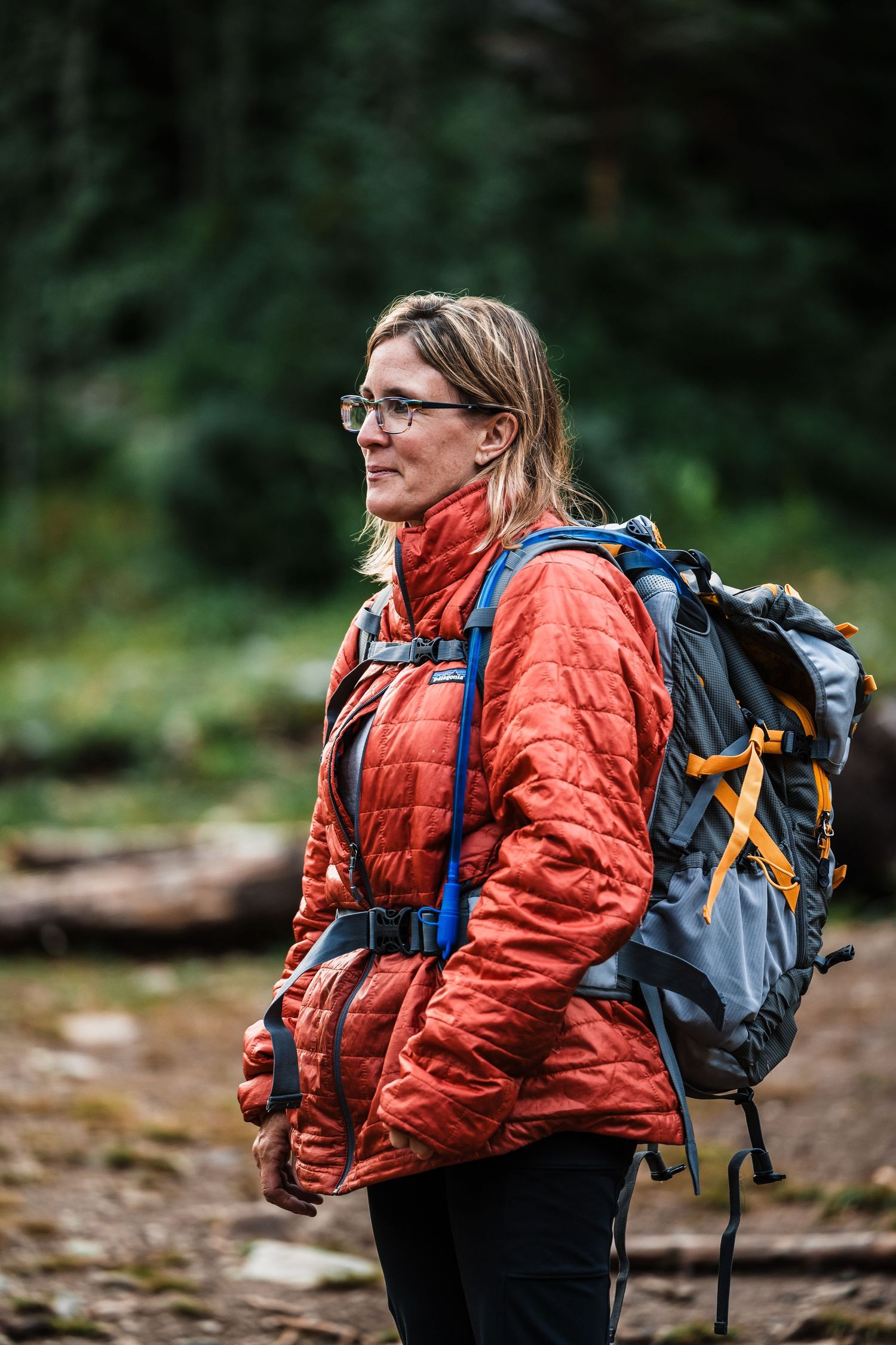 Hiker wearing orange puffy jacket and backpack pauses on forest trail.