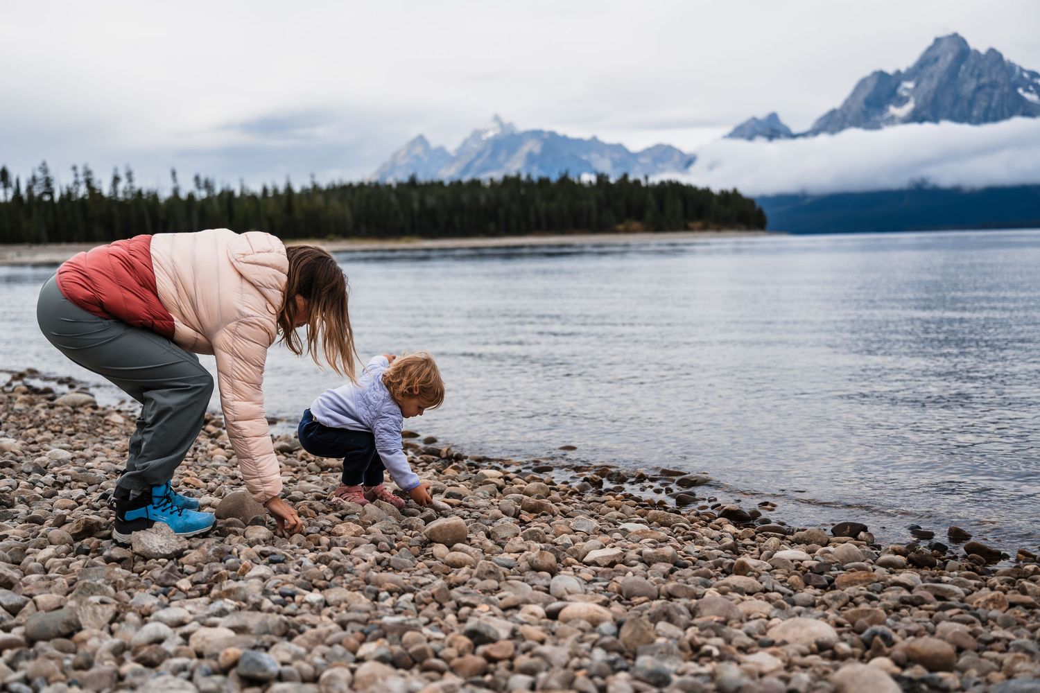 Person searching for rocks and shells along a scenic mountain lake shoreline at sunset.