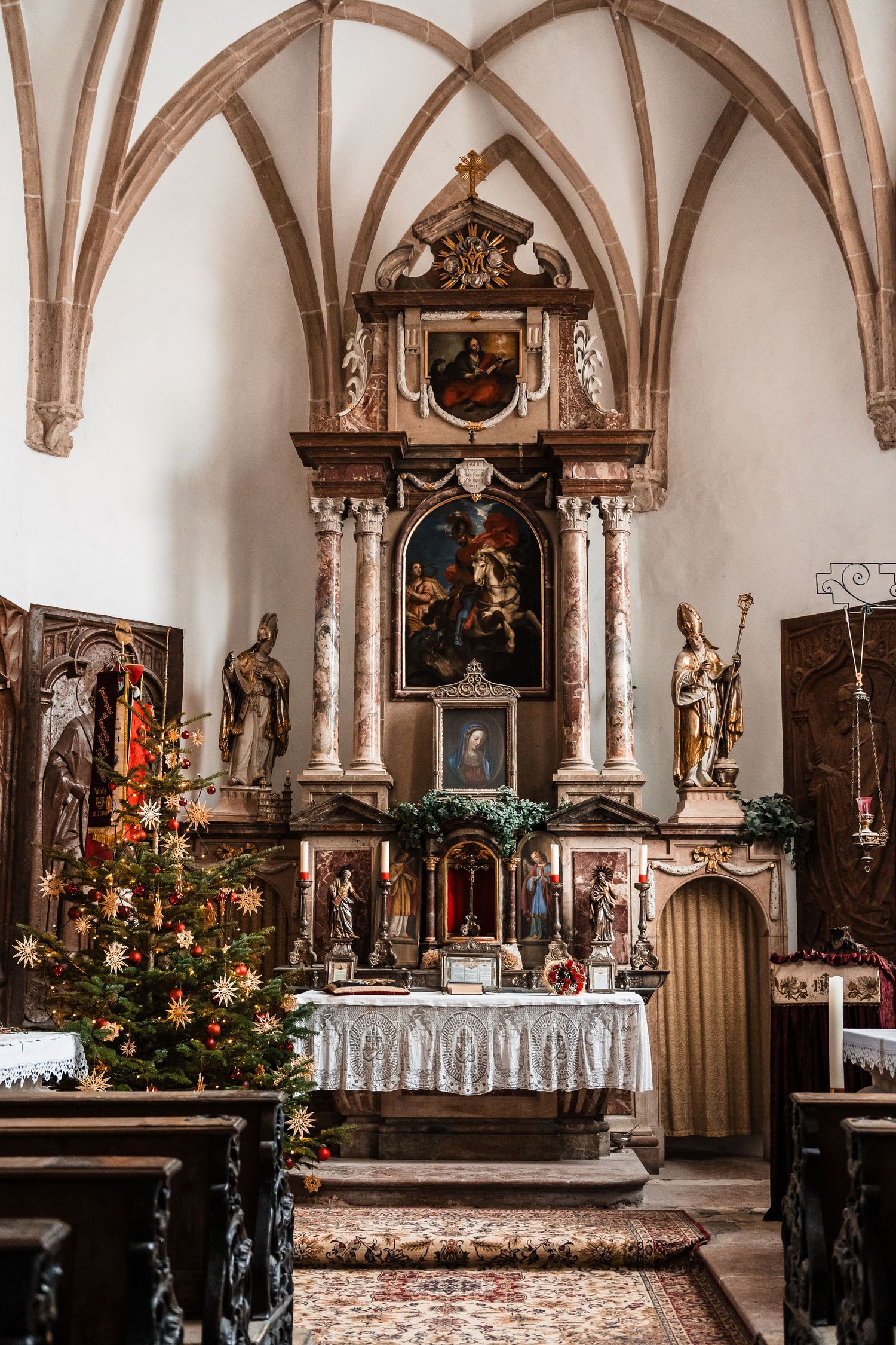 Gothic church interior with vaulted arches, religious statues, and decorated Christmas trees near the altar.