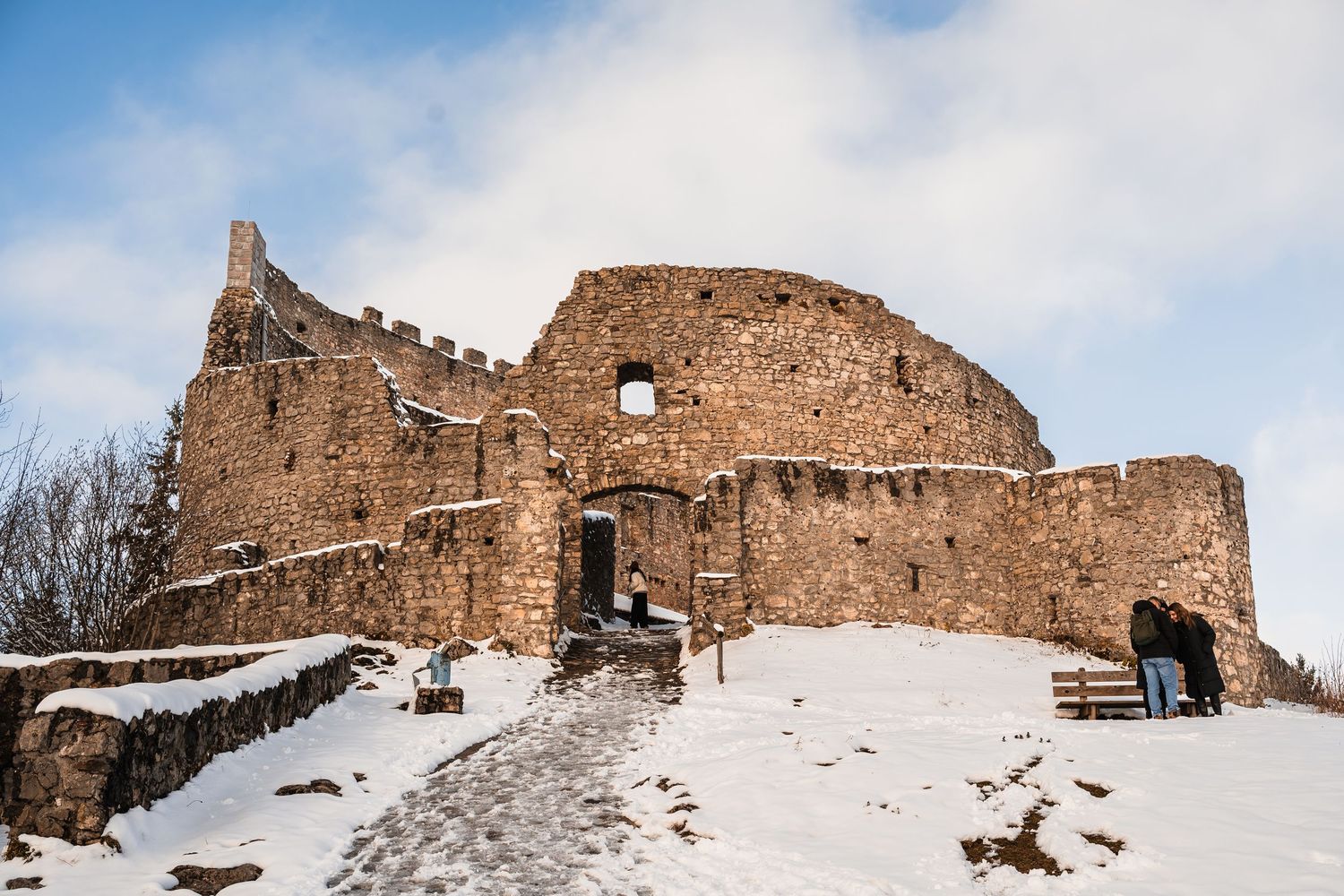 Snow-covered ruins of an ancient stone castle with crumbling walls against a cloudy winter sky.