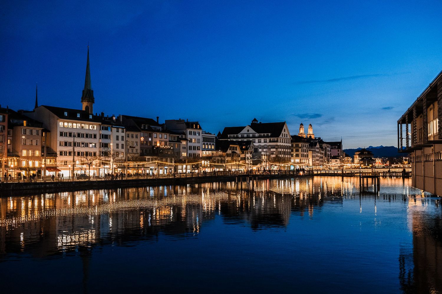 Panoramic twilight view of illuminated buildings and boats along Hamburg's historic harbor waterfront reflecting in calm water.