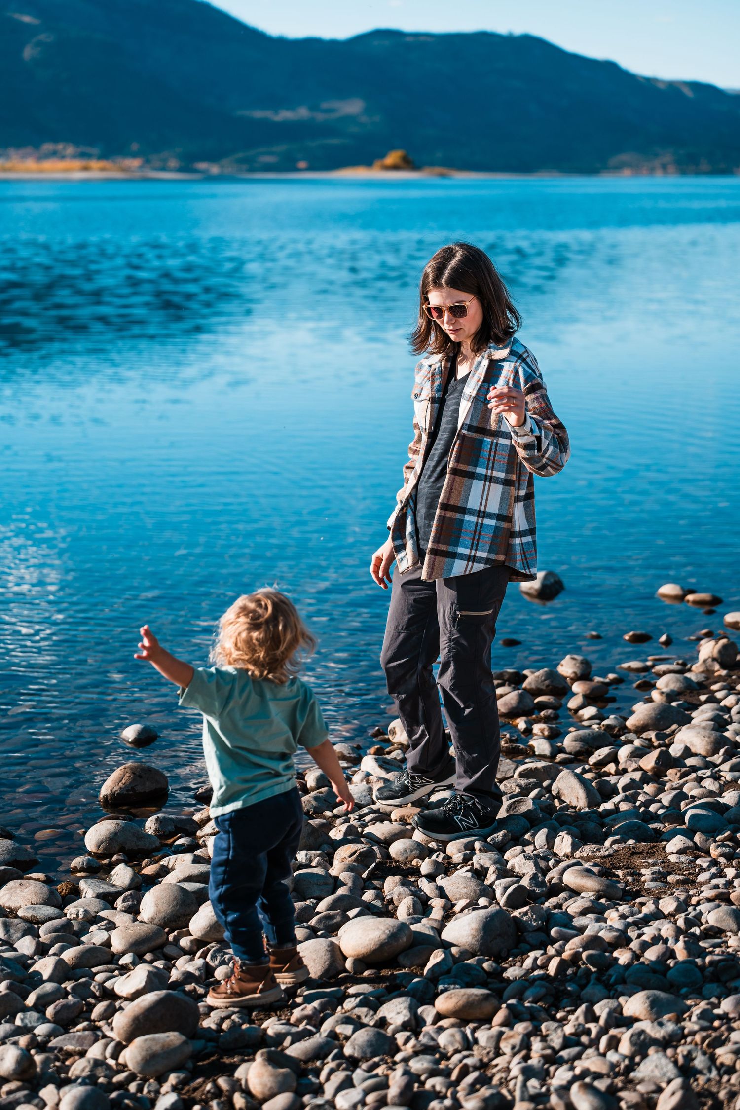 People exploring a rocky shoreline along a bright blue mountain lake on a sunny day.