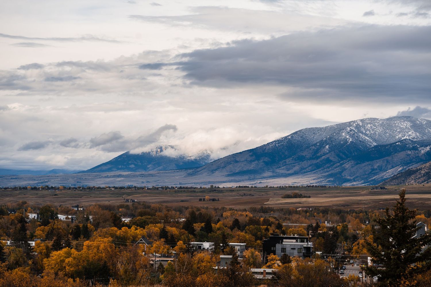 A panoramic view of snow-capped mountains and rolling hills under a dramatic cloudy sky in rural Montana.