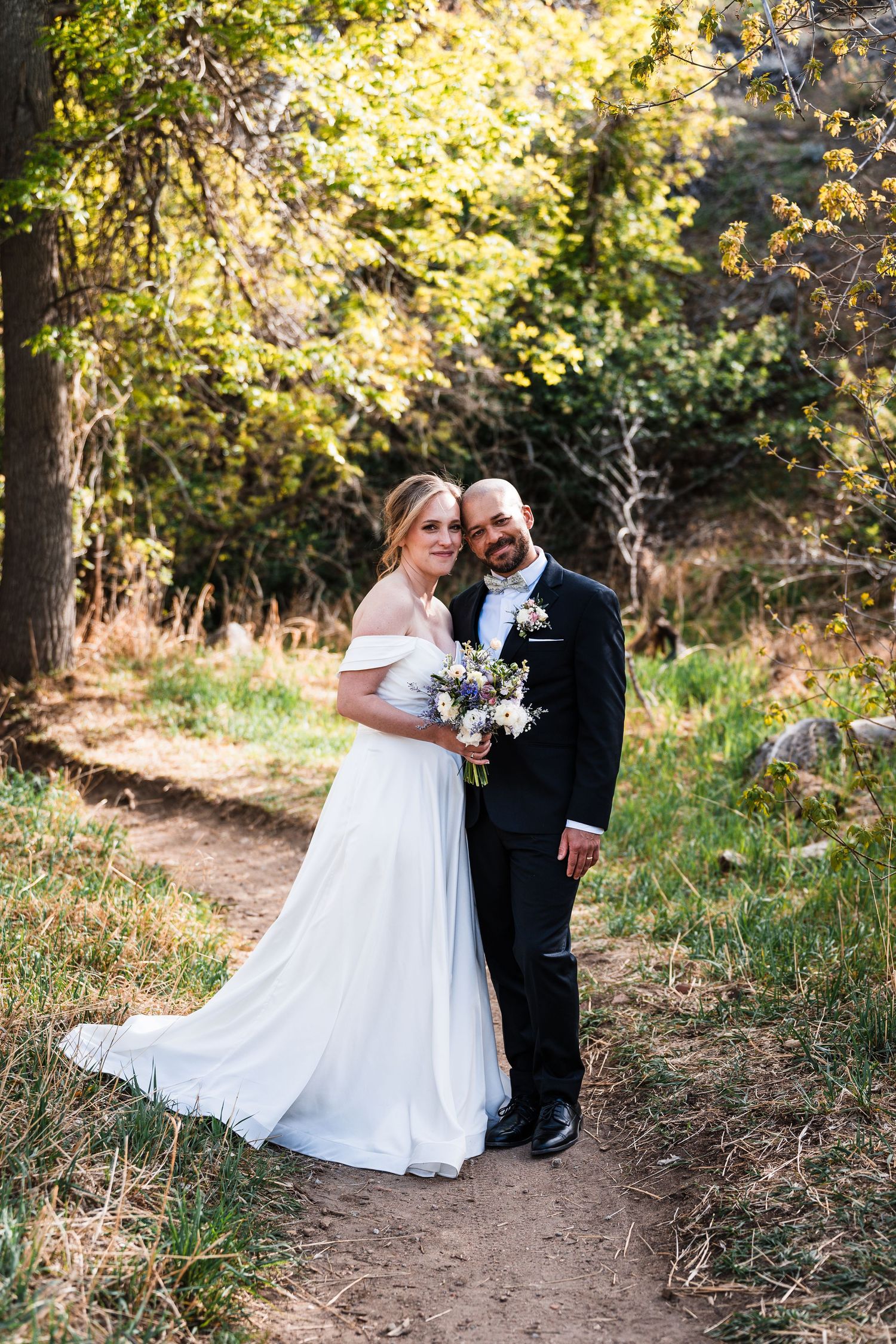 Bride and groom pose together on a wooded nature path holding a wildflower bouquet.