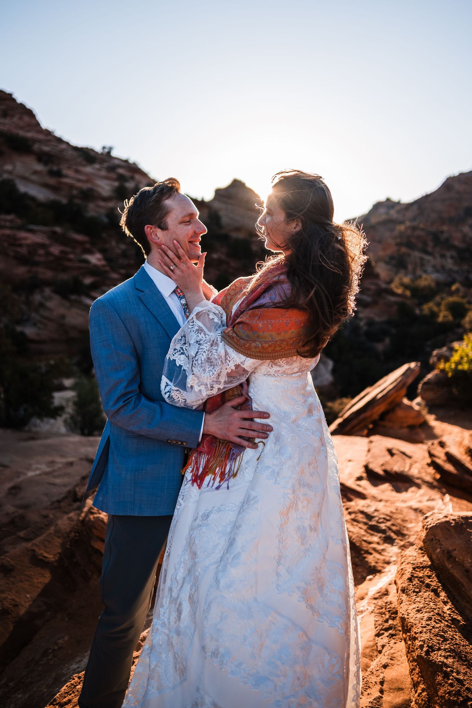 Bride and groom embrace at sunset among red rock formations, bride in lace dress with colorful shawl, groom in blue suit.