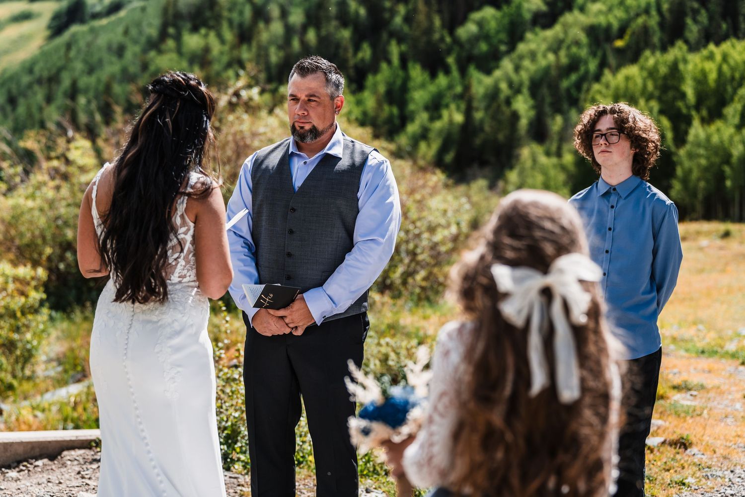 Groom in vest exchanges vows with bride outdoors during mountain wedding ceremony with guests watching.