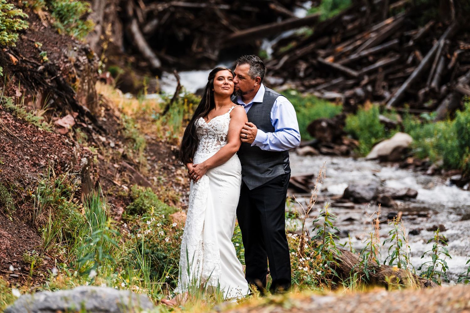 Romantic bride and groom portrait beside a rushing mountain stream surrounded by lush Colorado wilderness.