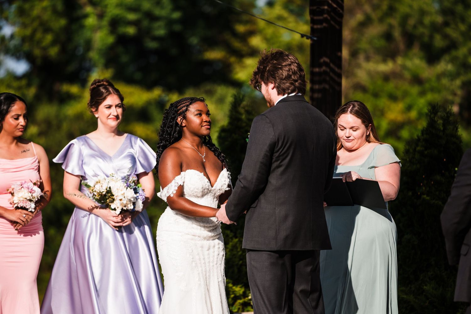 Bride and groom exchanging vows outdoors with bridesmaids in lavender and sage green dresses.