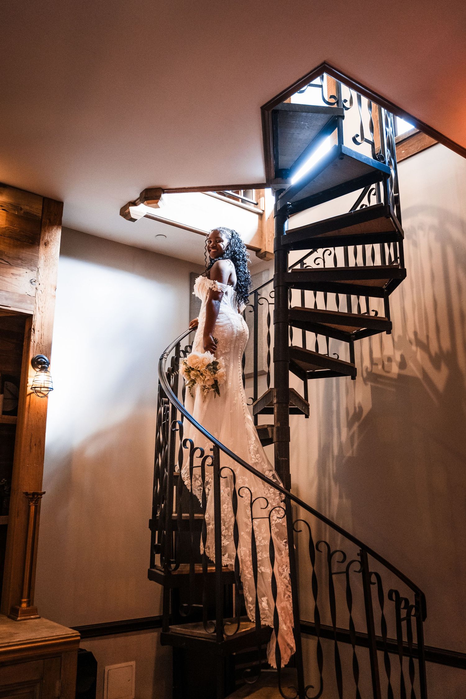 Elegant bride in lace gown ascending a spiral iron staircase holding a bouquet indoors.