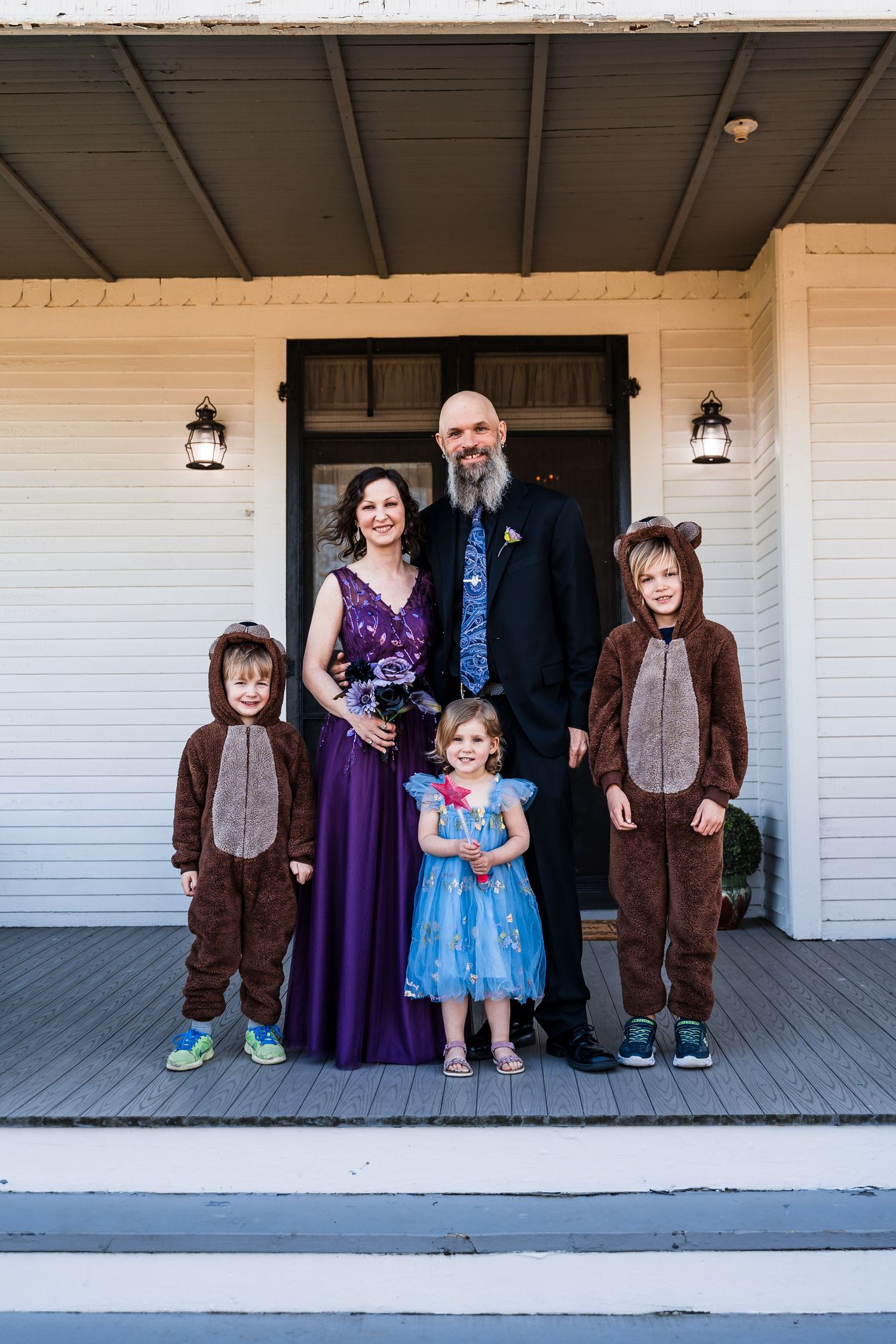 Family portrait with bald bearded groom, bride in purple gown, and three children in bear costumes on porch.