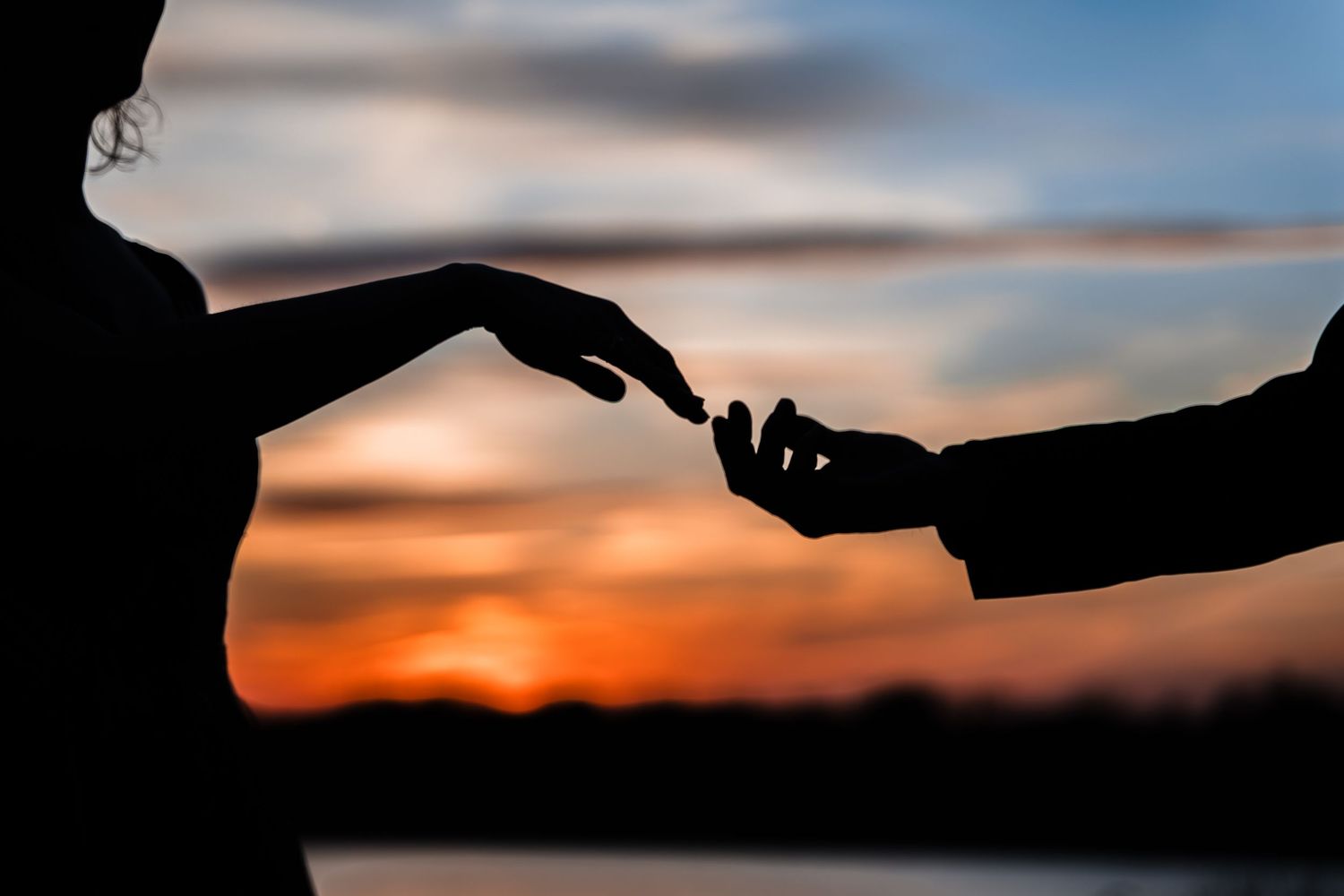 Silhouette of two people touching fingertips at sunset with colorful sky backdrop.