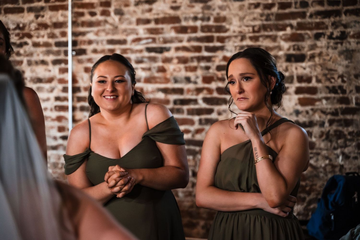 Women in olive green dresses share a candid moment against a rustic brick wall backdrop.