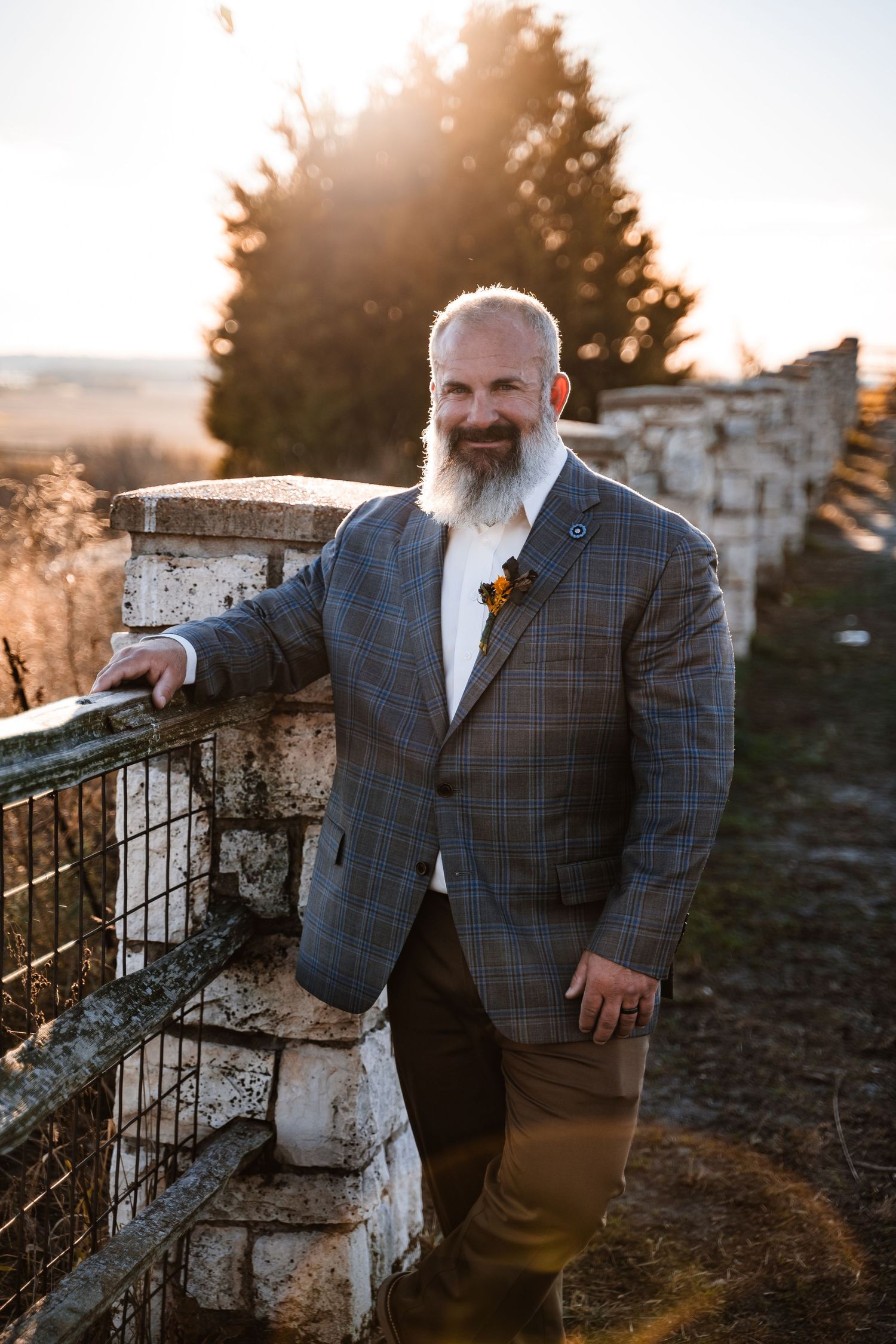A well-dressed person in a plaid suit jacket with a full white beard poses against a stone wall at sunset.