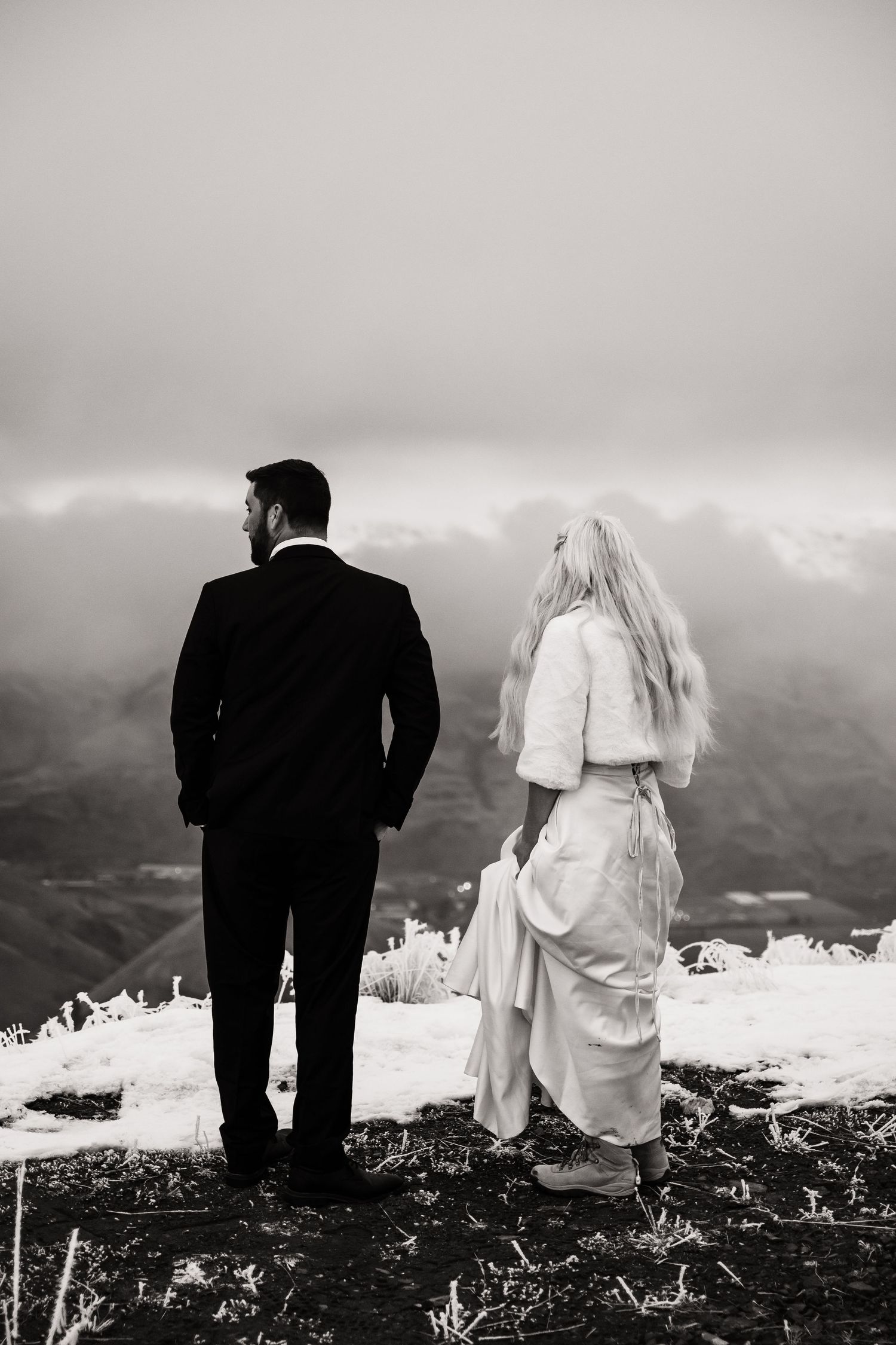 A black and white wedding photo of a couple standing on a snowy mountain overlook.