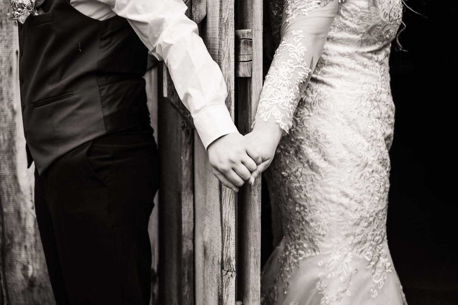 Black and white detail of bride and groom holding hands against rustic wooden wall.