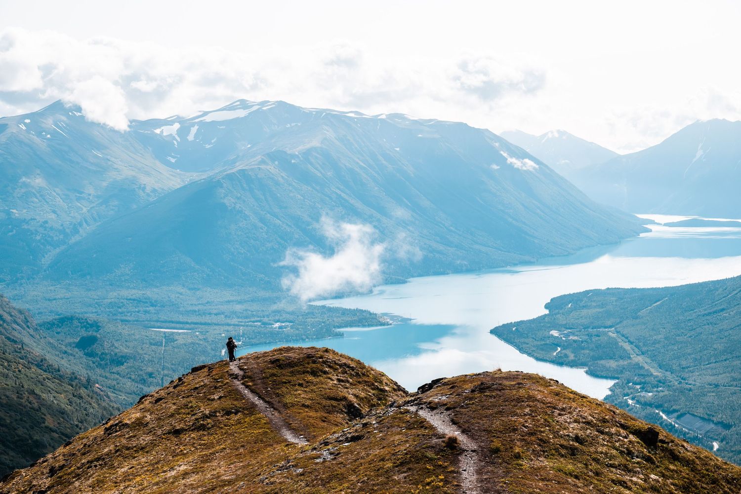 Lone hiker stands on mountain trail overlooking dramatic fjord and mountain ranges.