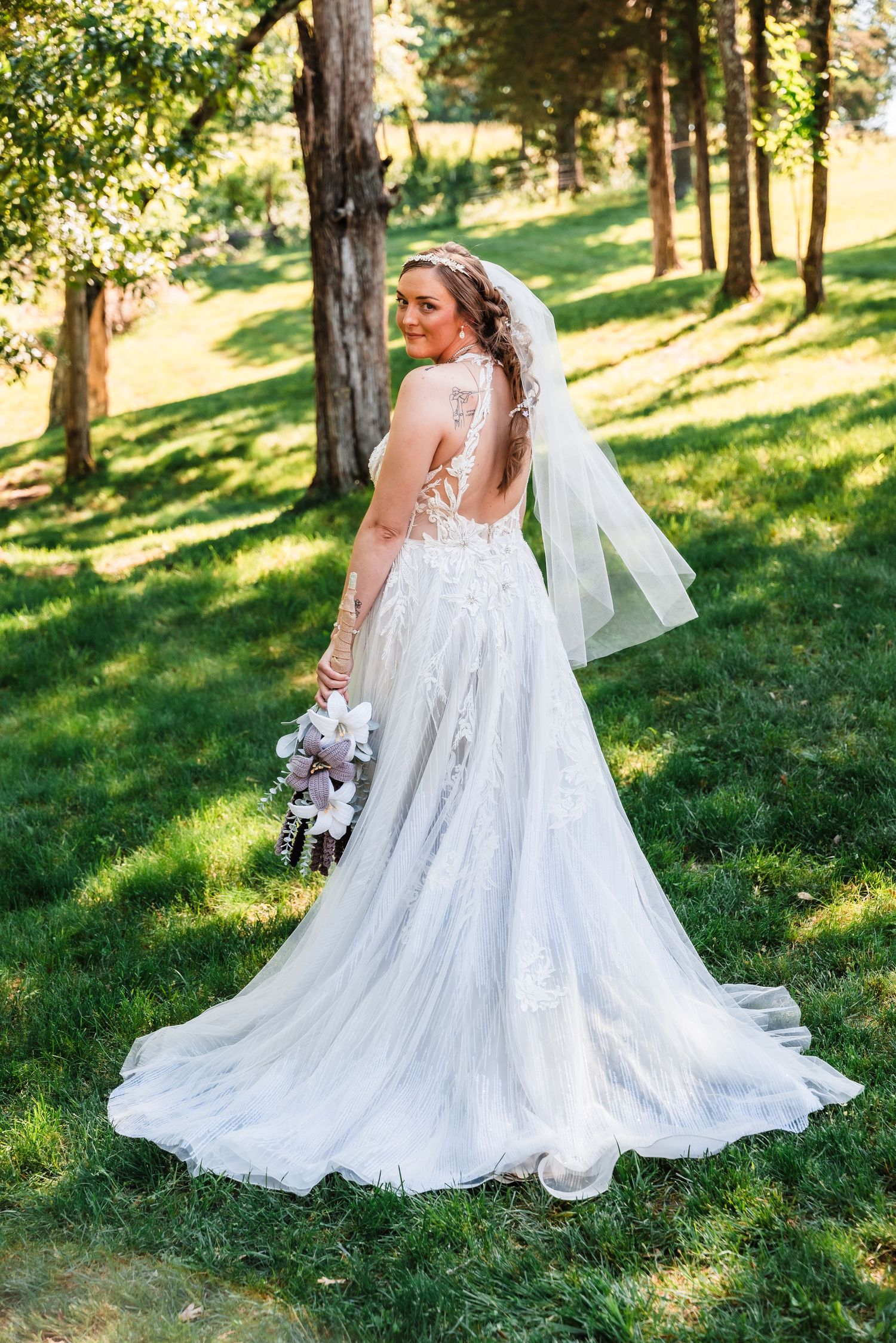 A bride in an elegant lace wedding gown with an open back poses in a sunlit wooded setting while holding a bouquet.