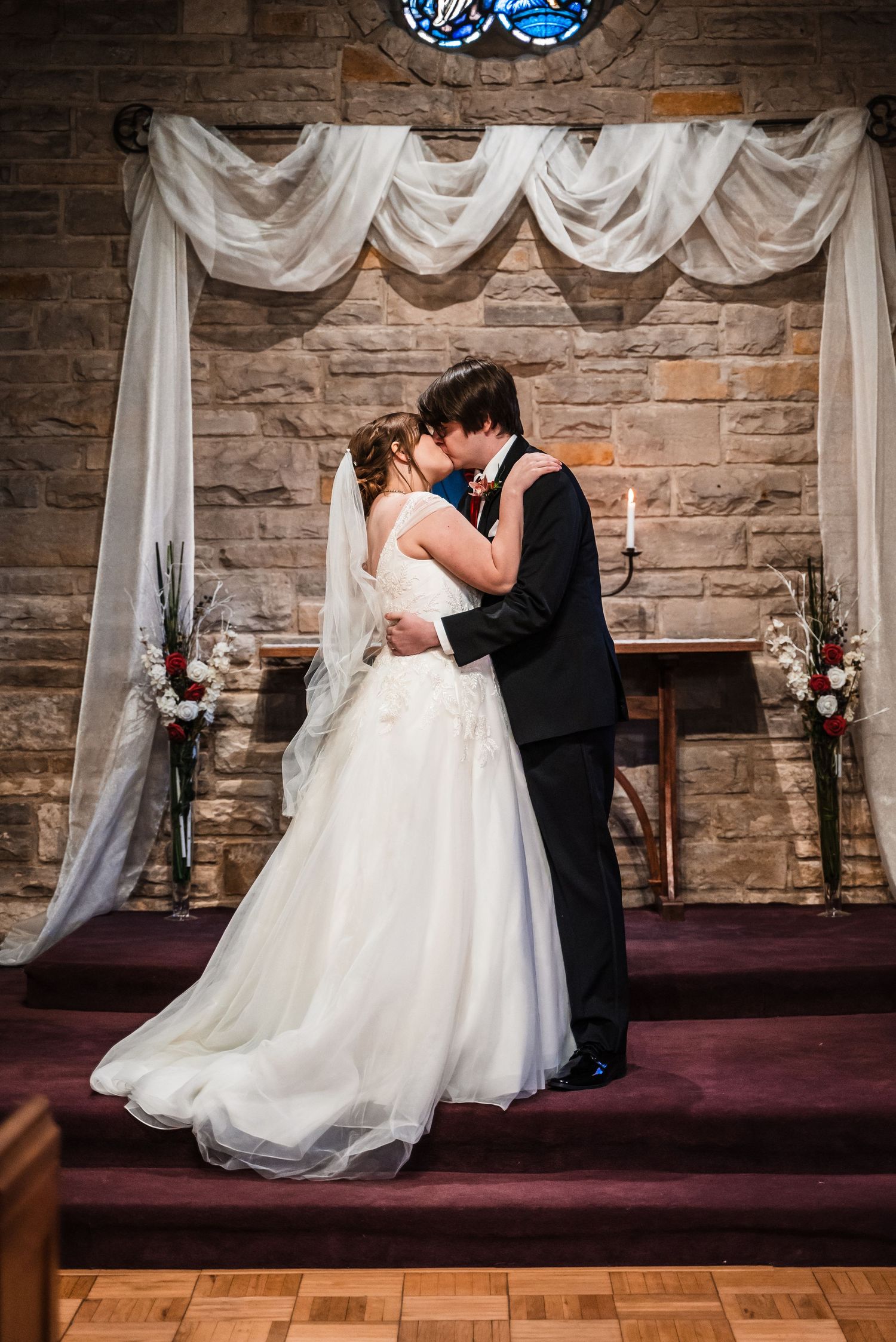 A romantic wedding kiss under draped white fabric and stone archway in a chapel setting.