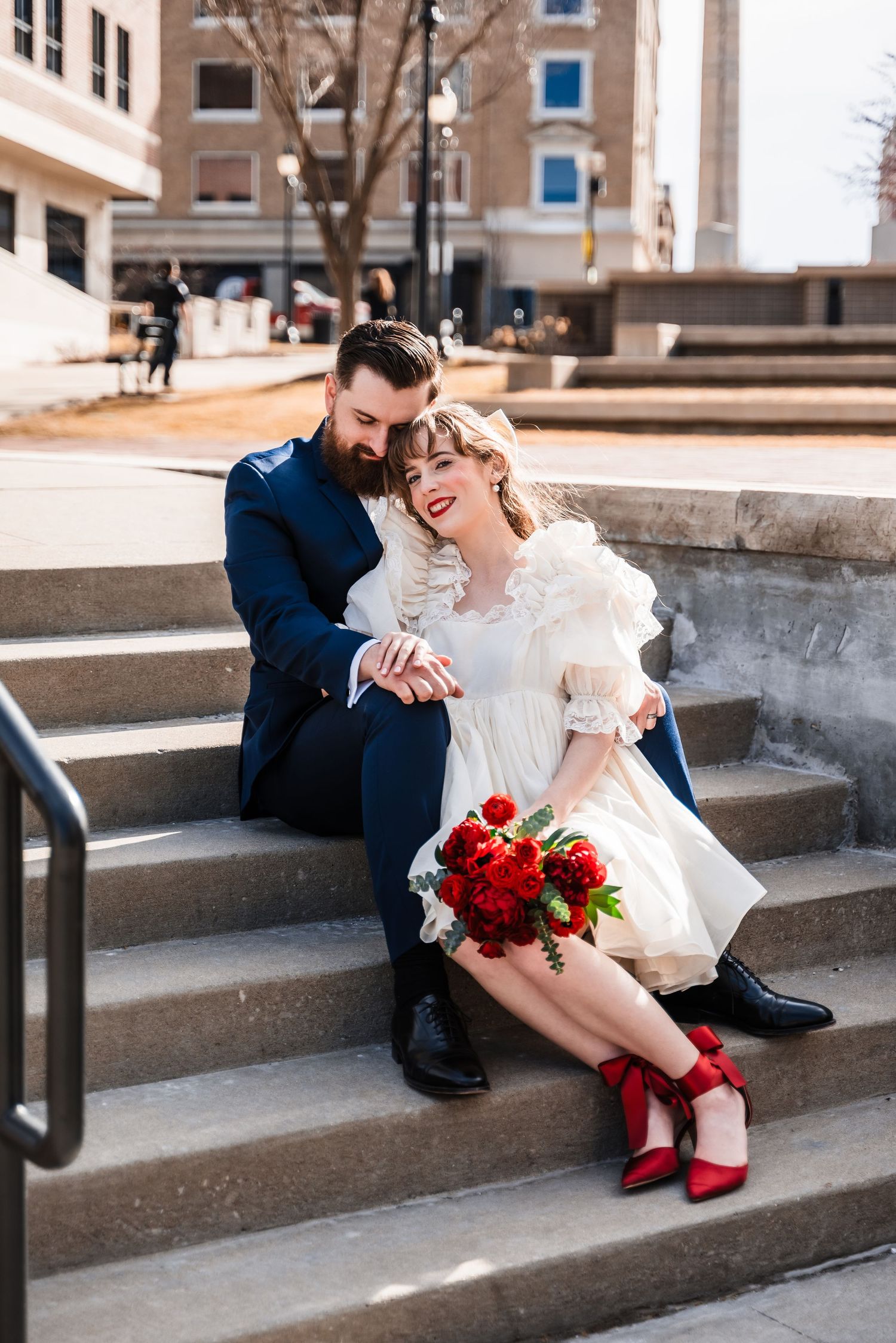 A couple sits on concrete steps sharing a tender moment with red roses and matching red shoes.