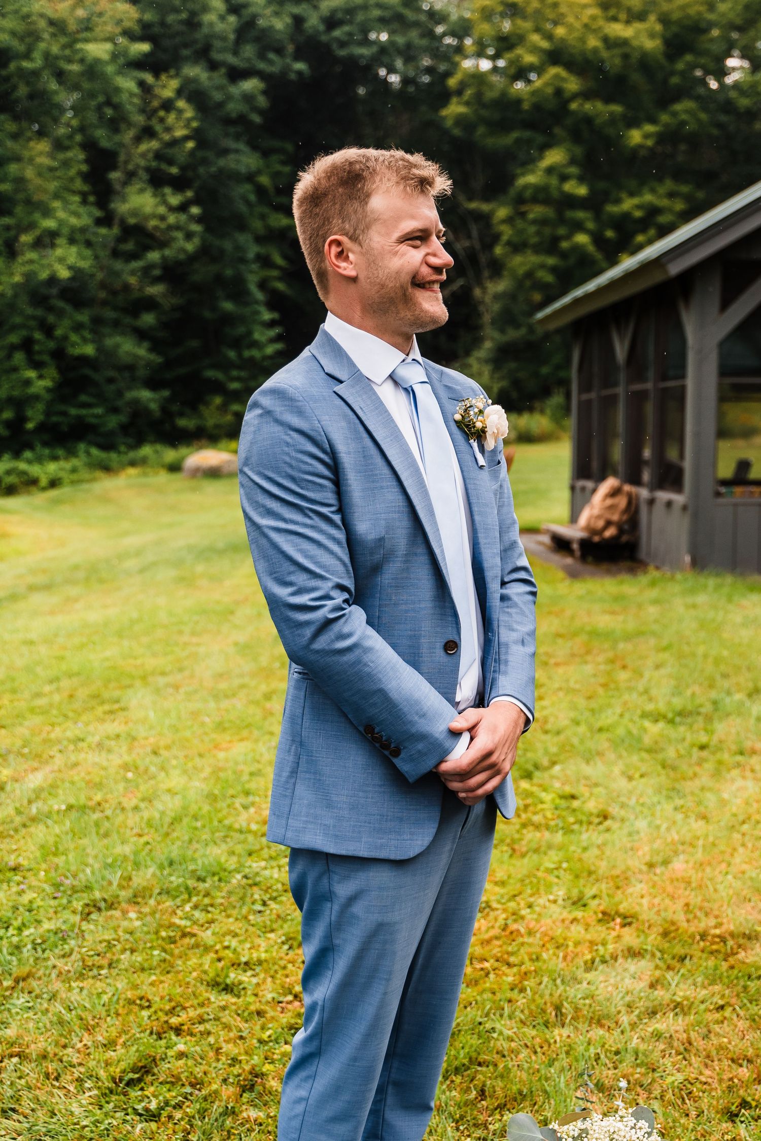 A smiling groom in a light blue suit stands on a grassy lawn during an outdoor wedding ceremony.