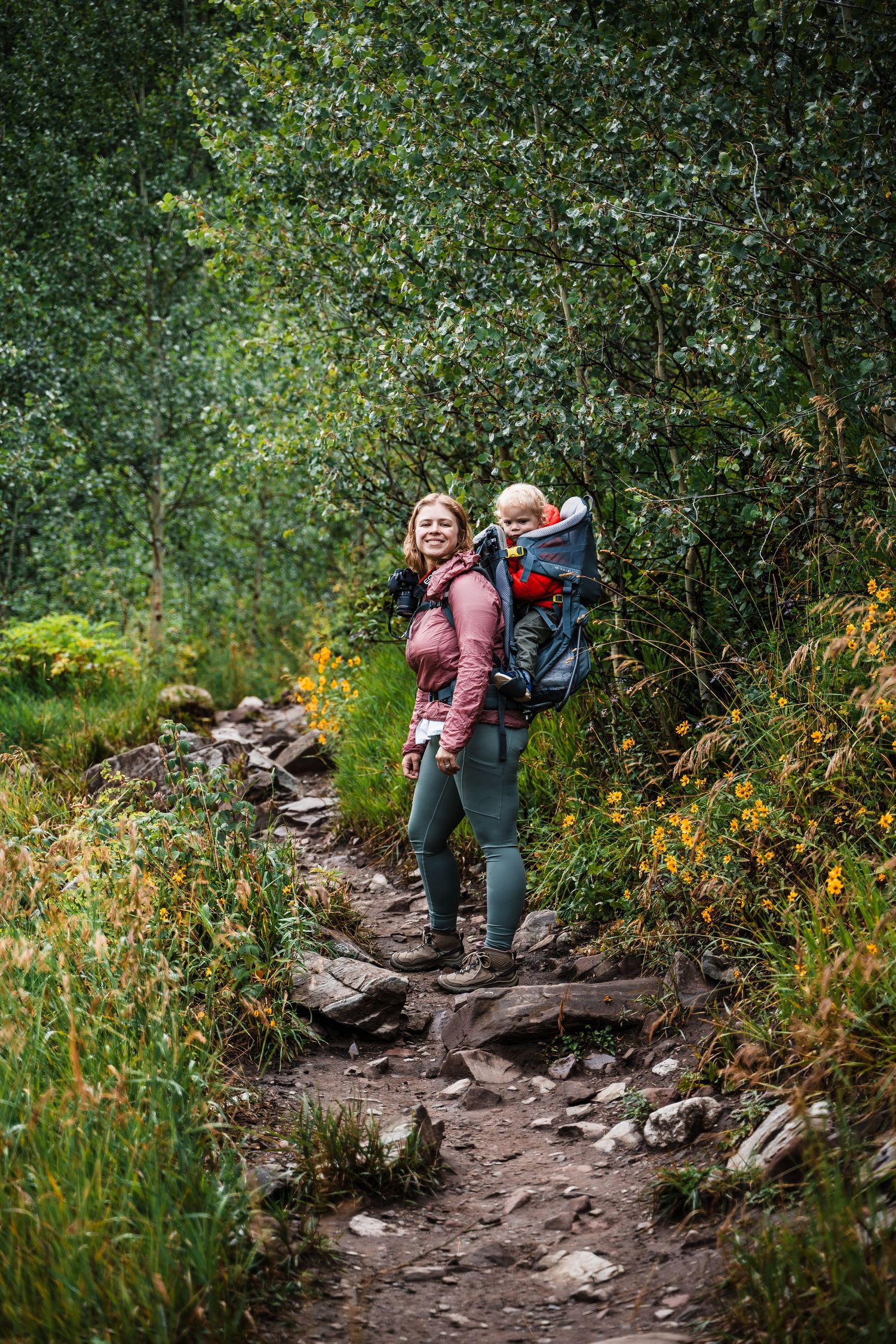 Hiker carrying child in backpack carrier walks along rocky mountain trail surrounded by greenery.