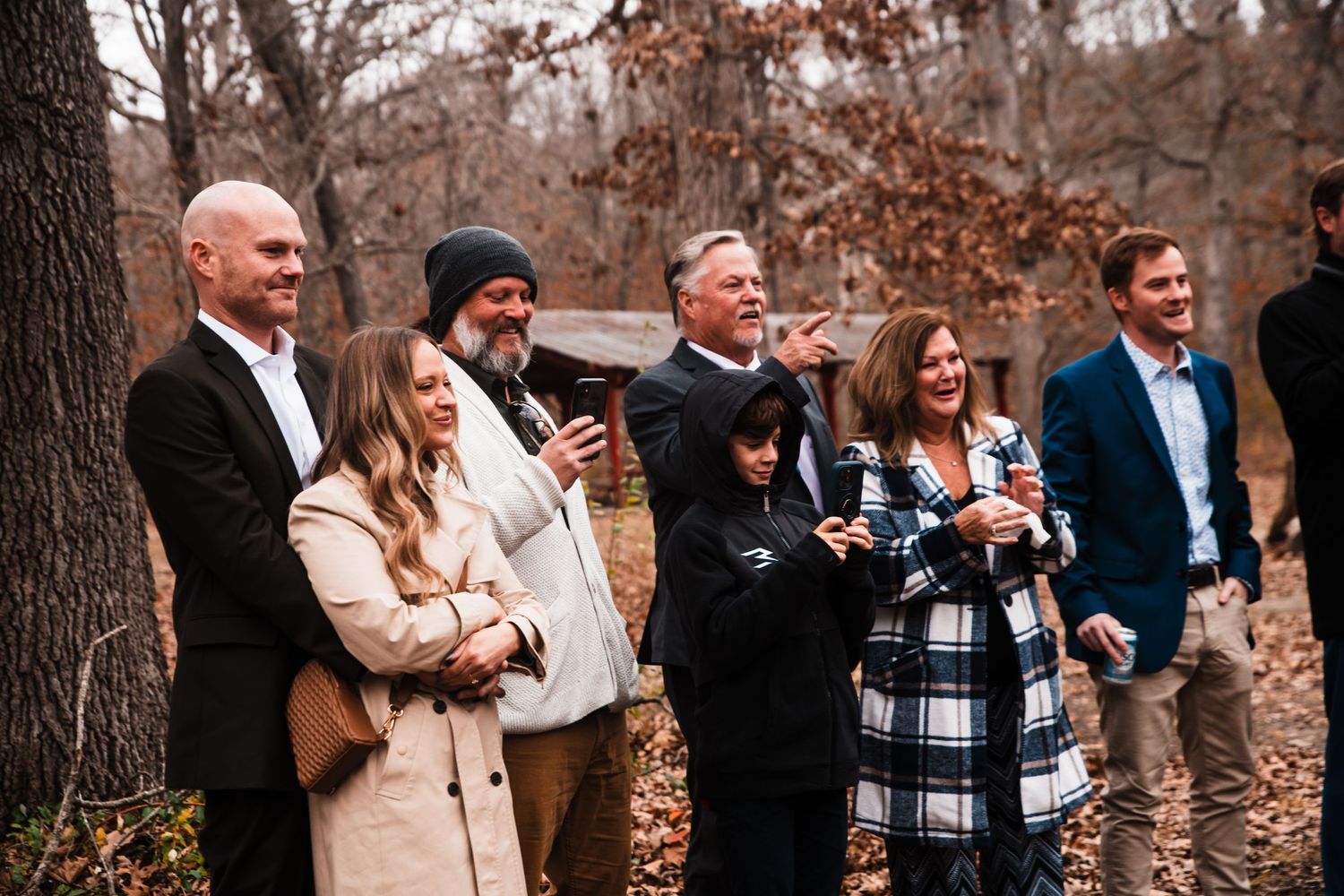 Wedding guests watching ceremony and smiling while standing among fall trees.