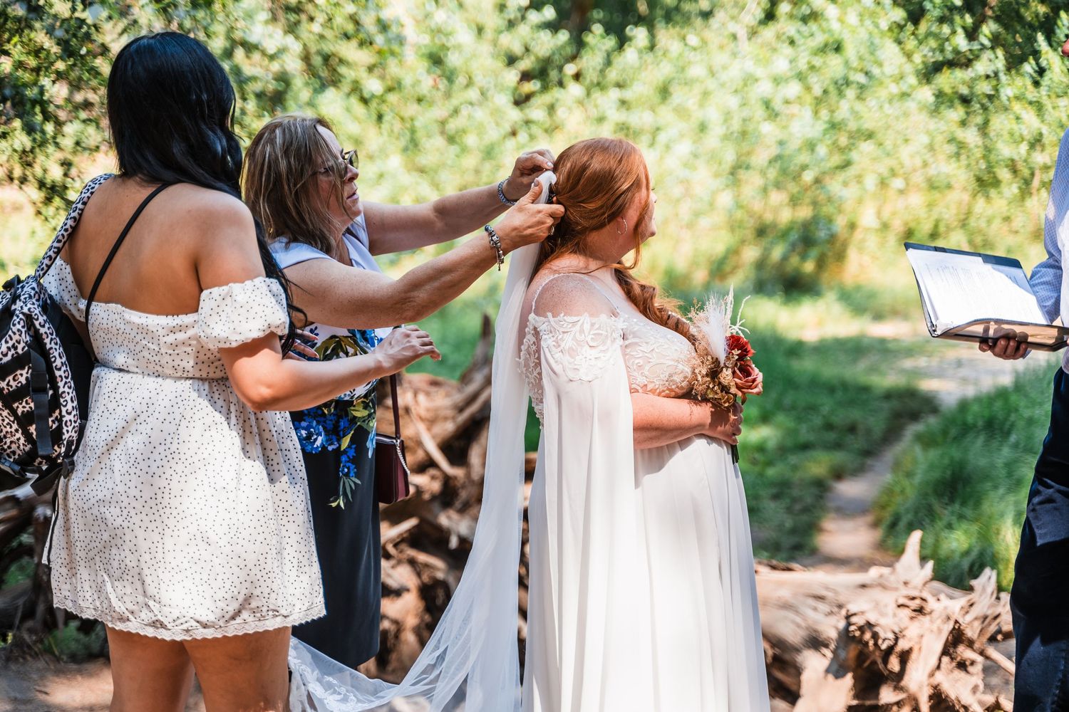 Bridesmaids in white lace dresses help with final preparations outdoors during a sunny wedding day.