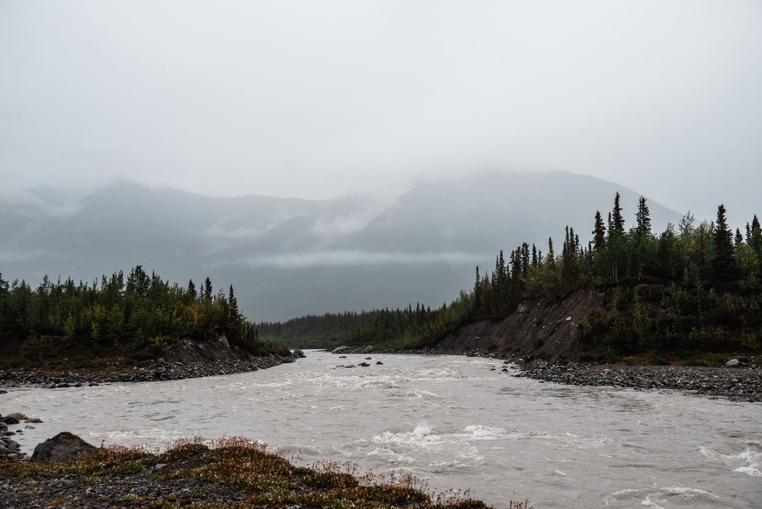 Turbulent river flows through a misty forested landscape with snow-capped mountains in the background.