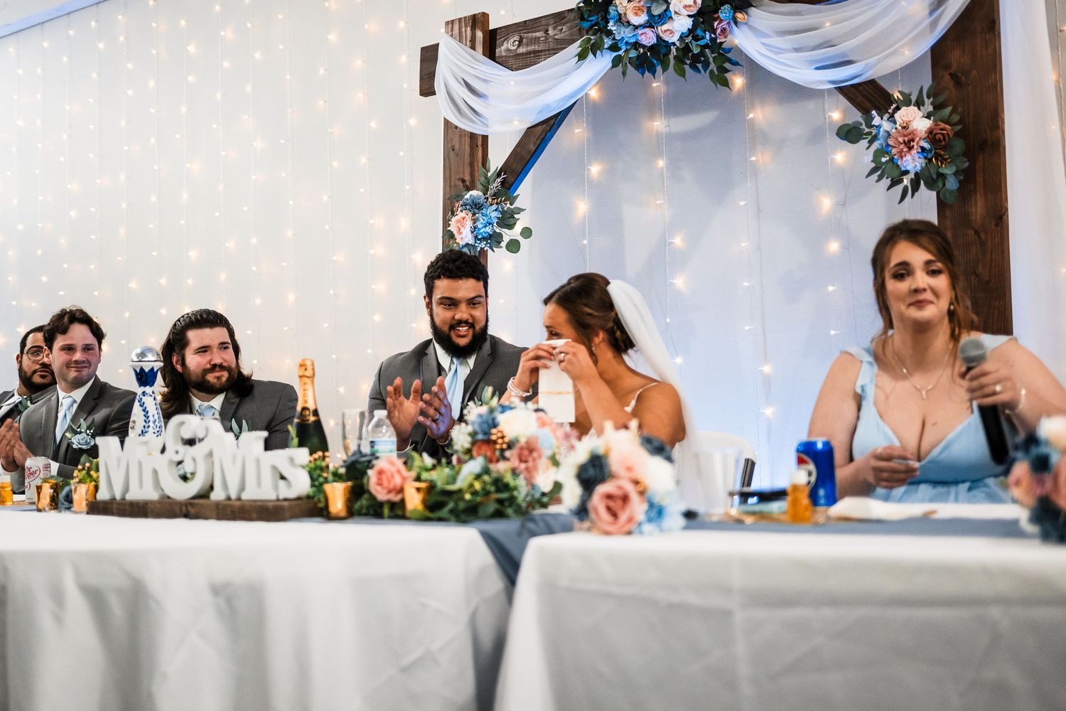 Wedding party laughs and celebrates at a reception table decorated with lights and draped fabric.