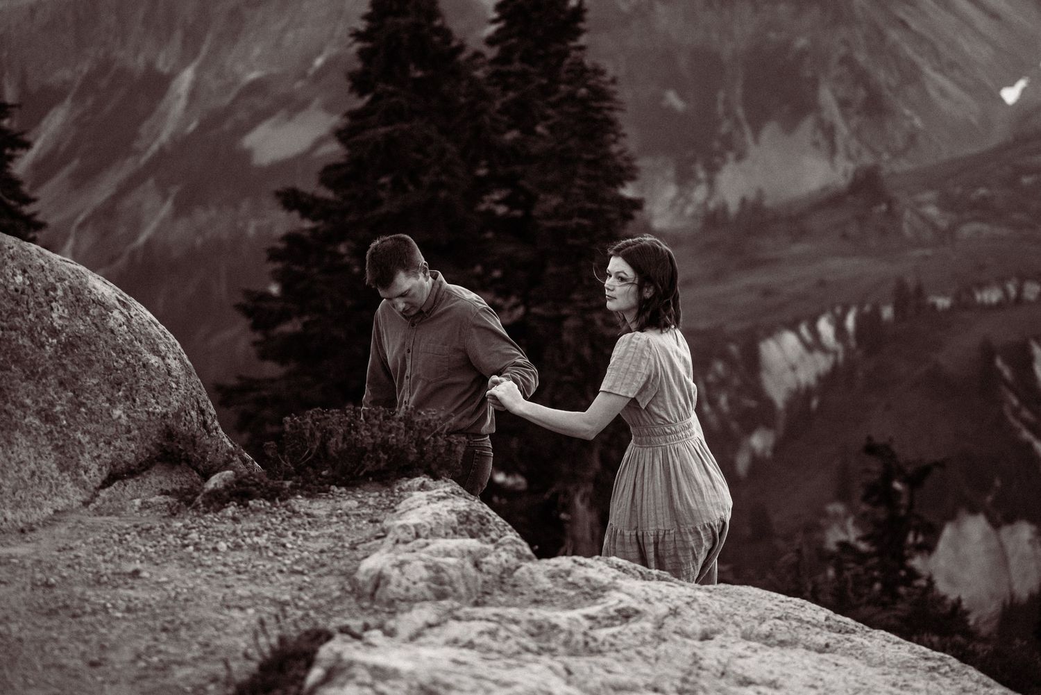 People sit on mountain ledge overlooking dramatic alpine landscape in black and white photograph.