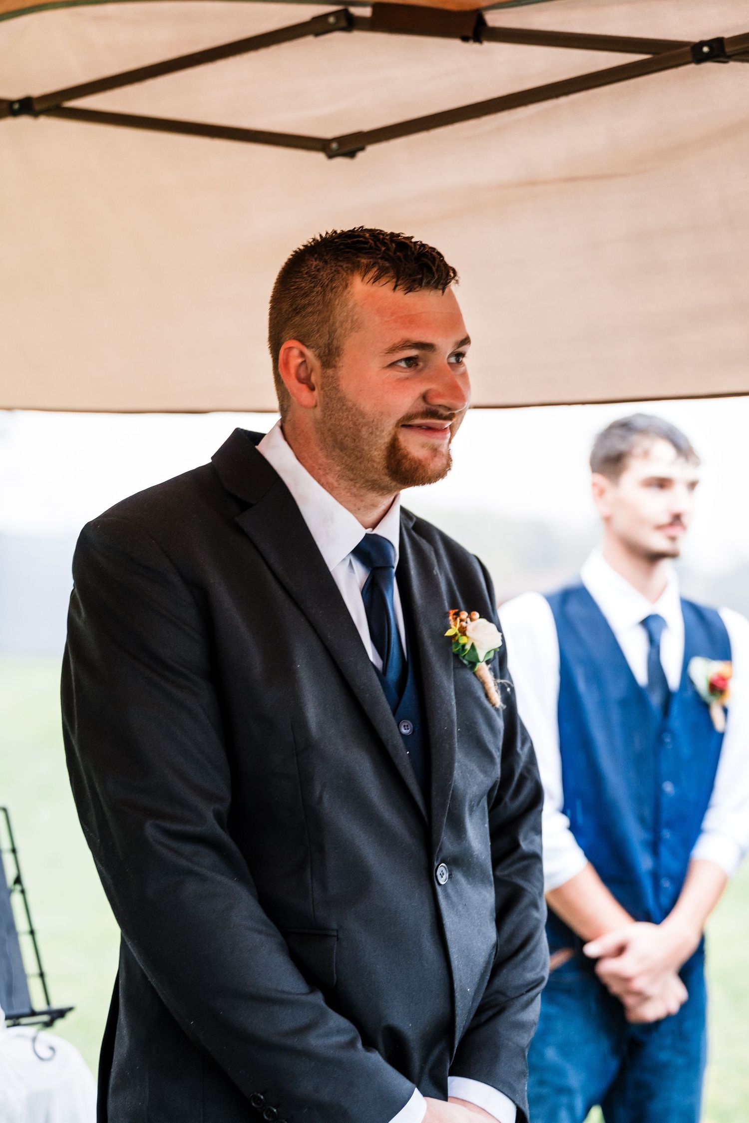 A row of groomsmen in dark suits and ties stand together under a white tent at an outdoor wedding.