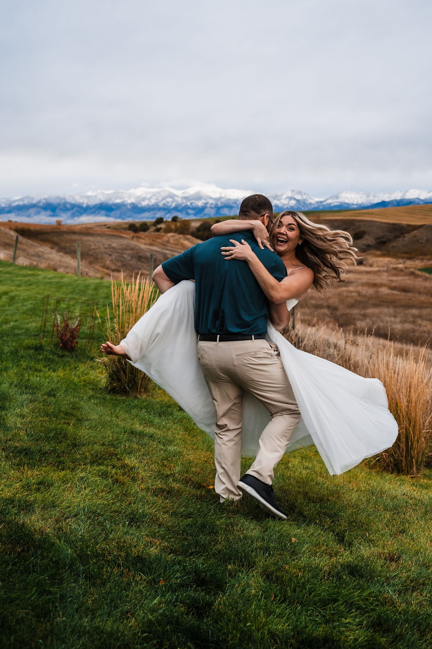 A couple in wedding attire share a romantic dip pose against a scenic mountain landscape backdrop.