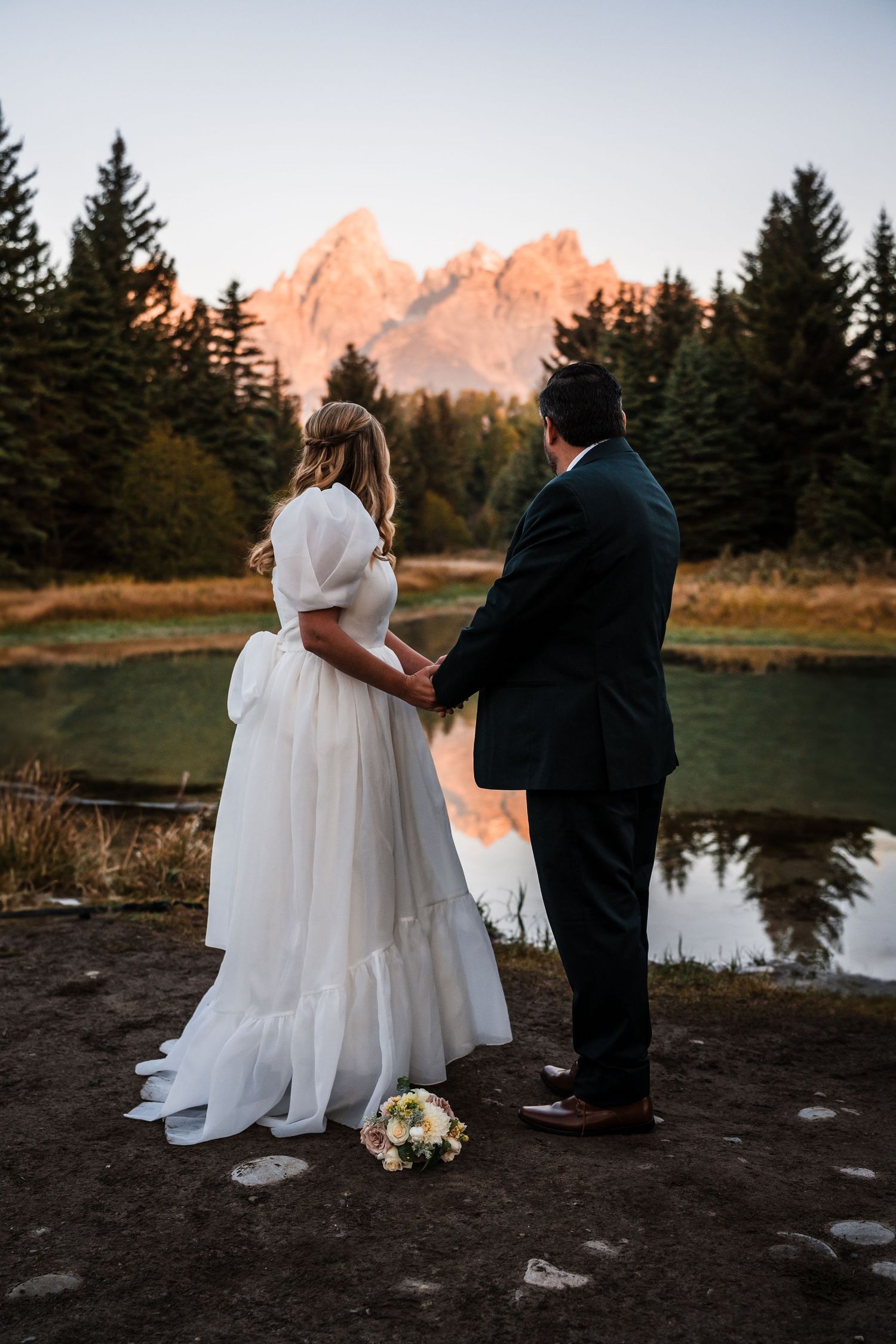 A couple in wedding attire stands by a lake at sunset with snow-capped mountains and evergreen trees in the background.