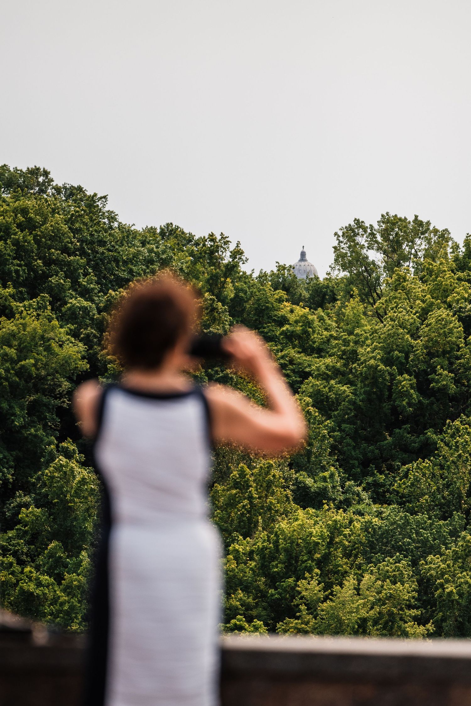 A figure in a white tank top gazes at a distant church spire through lush green trees on an overcast day.