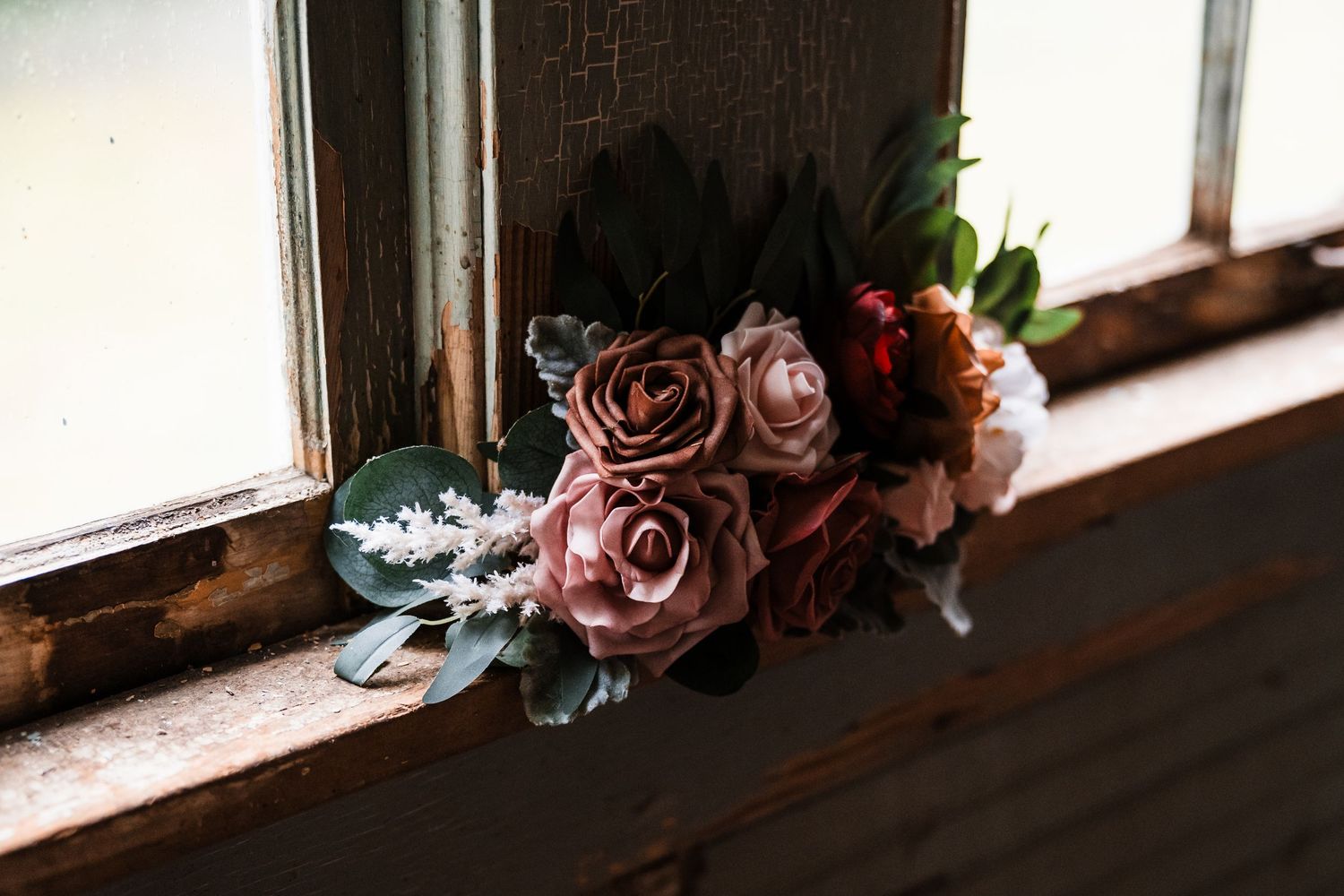 A vintage dusty pink rose bouquet with dark burgundy flowers and eucalyptus rests on an old wooden windowsill.
