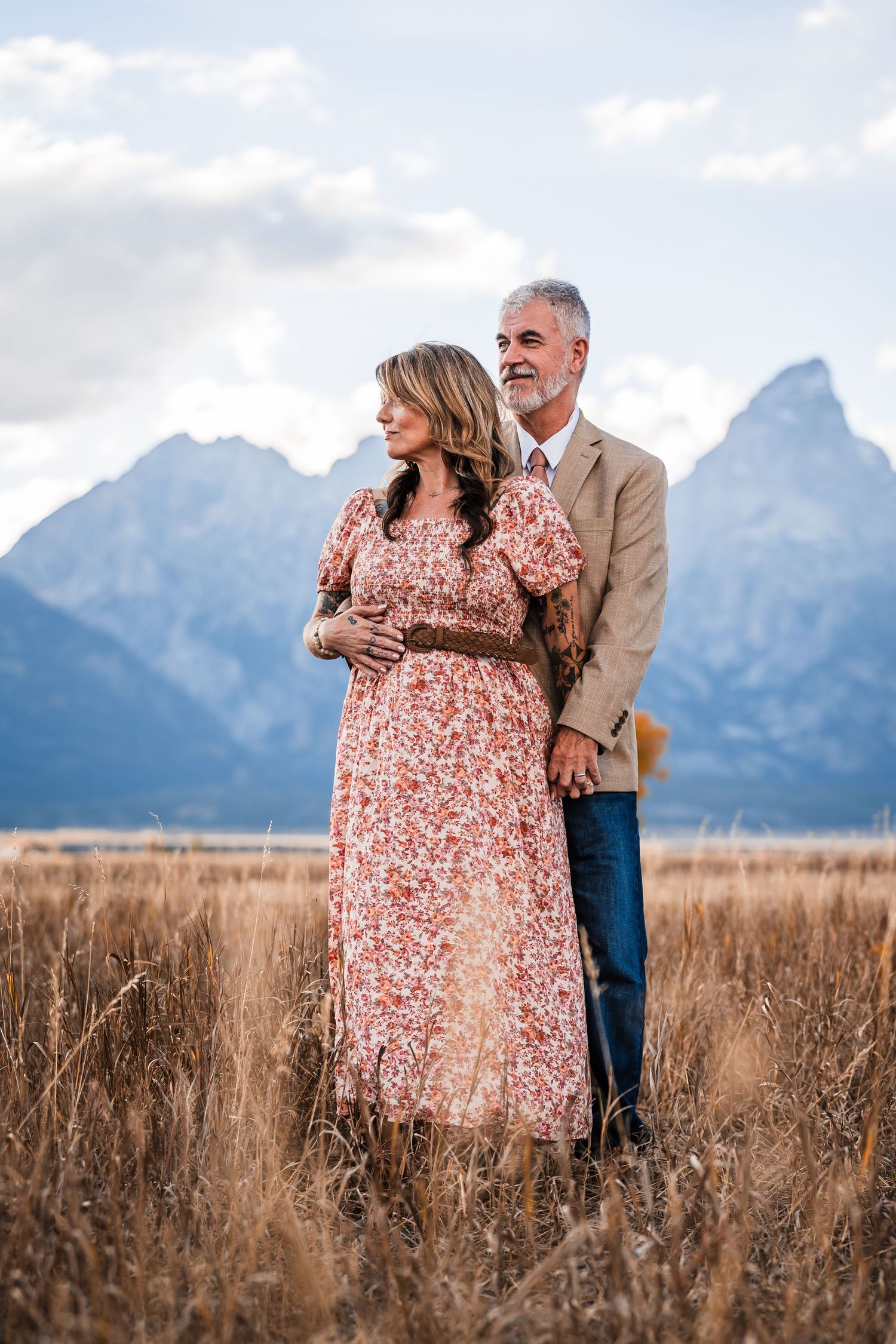 A couple embraces in a field with the majestic Grand Teton mountains rising dramatically in the background.